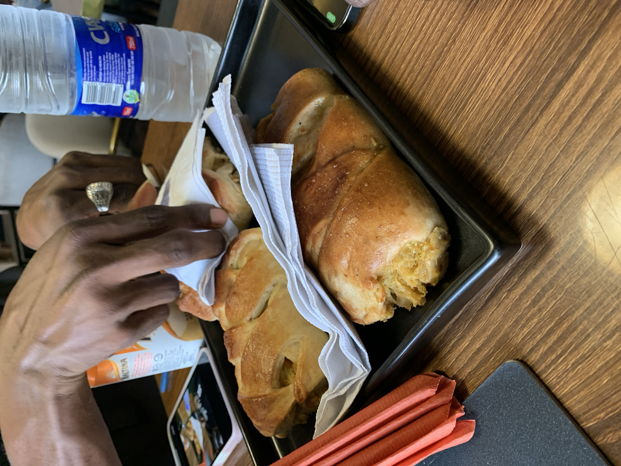 A tray containing two pastries wrapped in white napkins is shown on a wooden table. A person's hands are reaching towards one of the pastries. A bottle of water is visible in the background. A smartphone and a container with orange packaging are also on the table. Folded orange napkins are near the tray. The location is Adankolo, Nigeria.