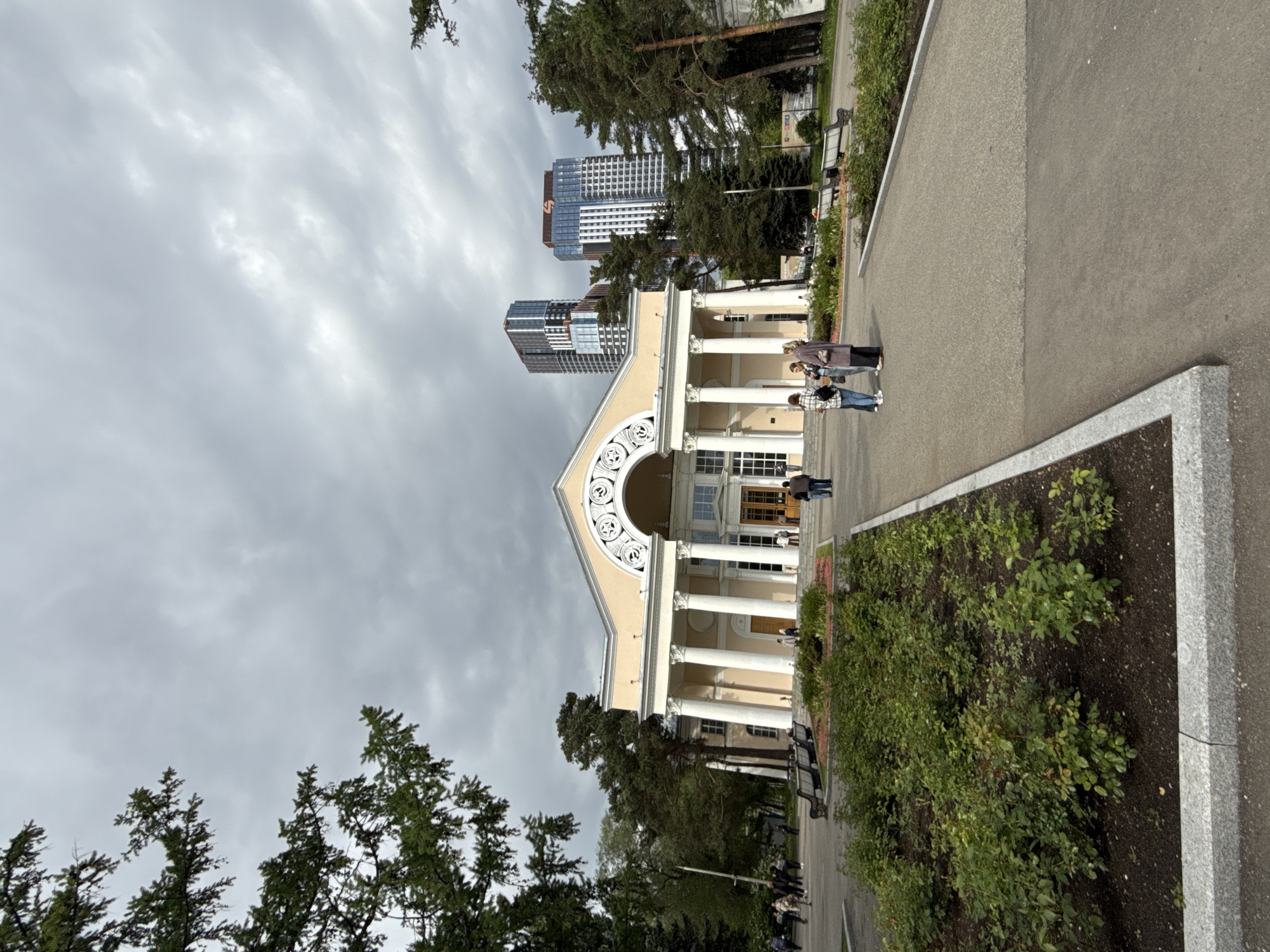 The image shows a building with tall columns in the Gorky Central Park in Moscow, Russia. The architecture of the building is classical in design, featuring off-white pillars and ornate detailing above the entrance. Modern buildings loom in the background behind the building. A wide paved area, likely a pathway, runs along the side of the building. Bushes and trees are interspersed along the pathway. Several people are seen walking along the pathway, with a group of about five pedestrians in the middle distance, and others further away. The sky is overcast, suggesting a cloudy or potentially rainy day.
