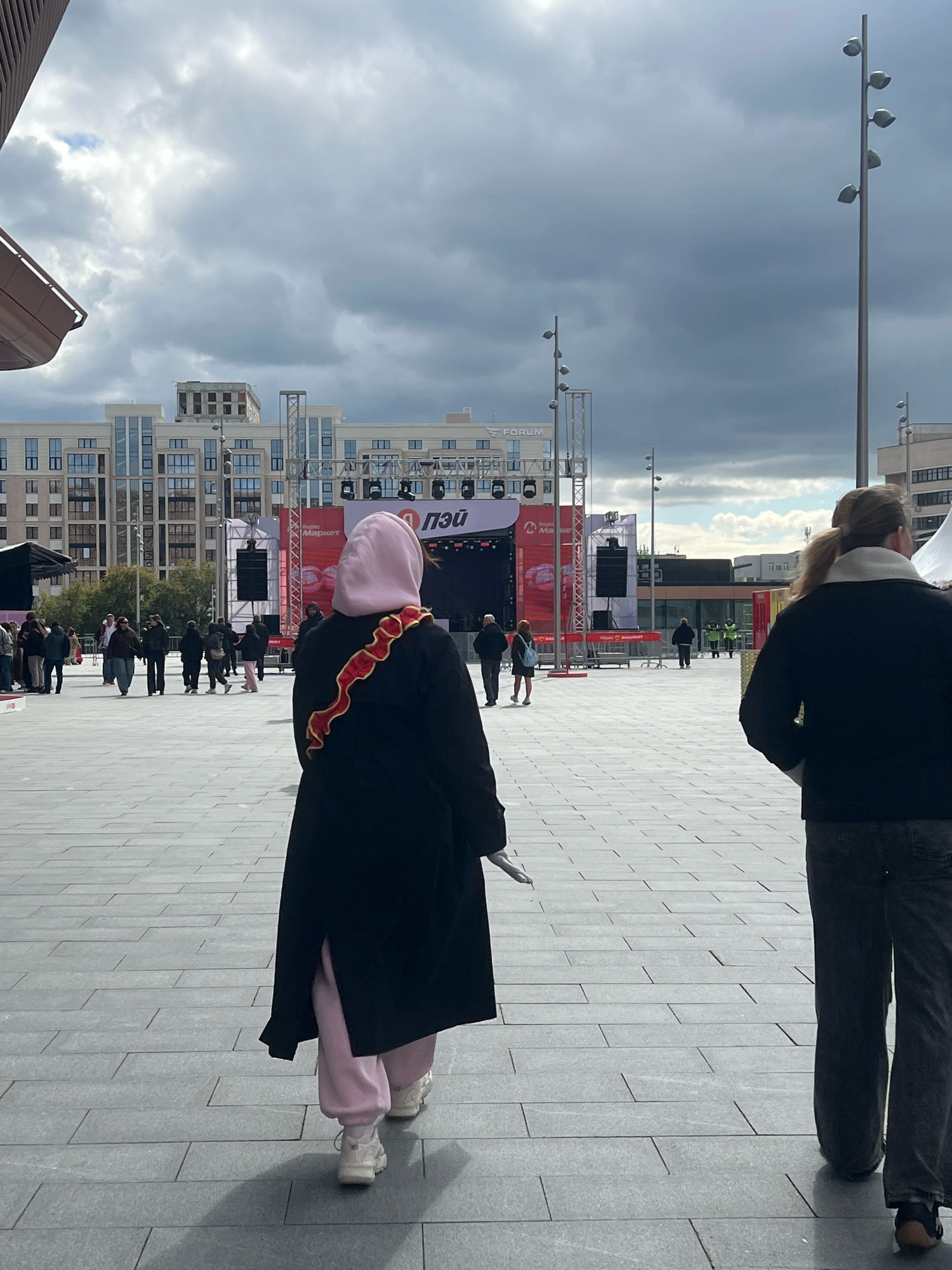 An outdoor daytime scene features a large, paved public square in front of a stage setup. Several people are visible, including a woman in the foreground wearing a black coat, a pink hooded garment, and light pink pants, with a red and yellow ribbon across her back. To her right, another person is partially visible from behind, wearing a dark jacket and dark pants. In the middle ground, a group of people are walking, and behind them is a stage with a large screen displaying Russian text and logos. In the background, modern multi-story buildings are visible. The sky is overcast with dramatic clouds. This appears to be the 1905 Square in Yekaterinburg, Russia, a common venue for public events and concerts, as suggested by the stage setup and building architecture which are consistent with landmarks in that area.