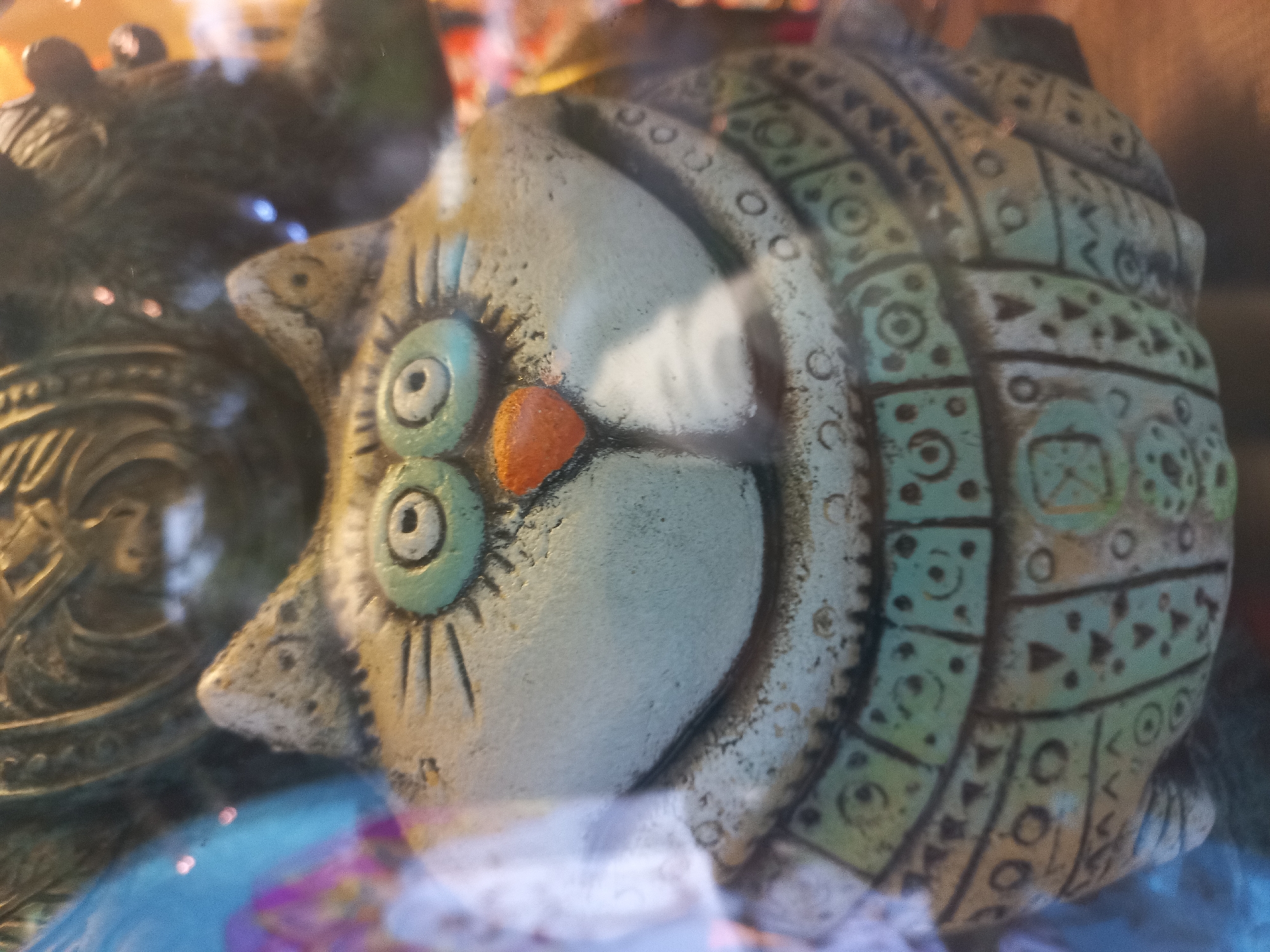 A close-up shot shows a ceramic owl figurine with large, round, light green eyes and an orange triangular nose. The figurine is decorated with geometric patterns in darker shades of green and brown. It is placed near another decorative object, possibly also ceramic, featuring a face. The setting includes hints of blue and purple colors, suggesting a possible indoor or display environment. There is no indication that the image was taken in Sochi, Russia, or any location in Russia.