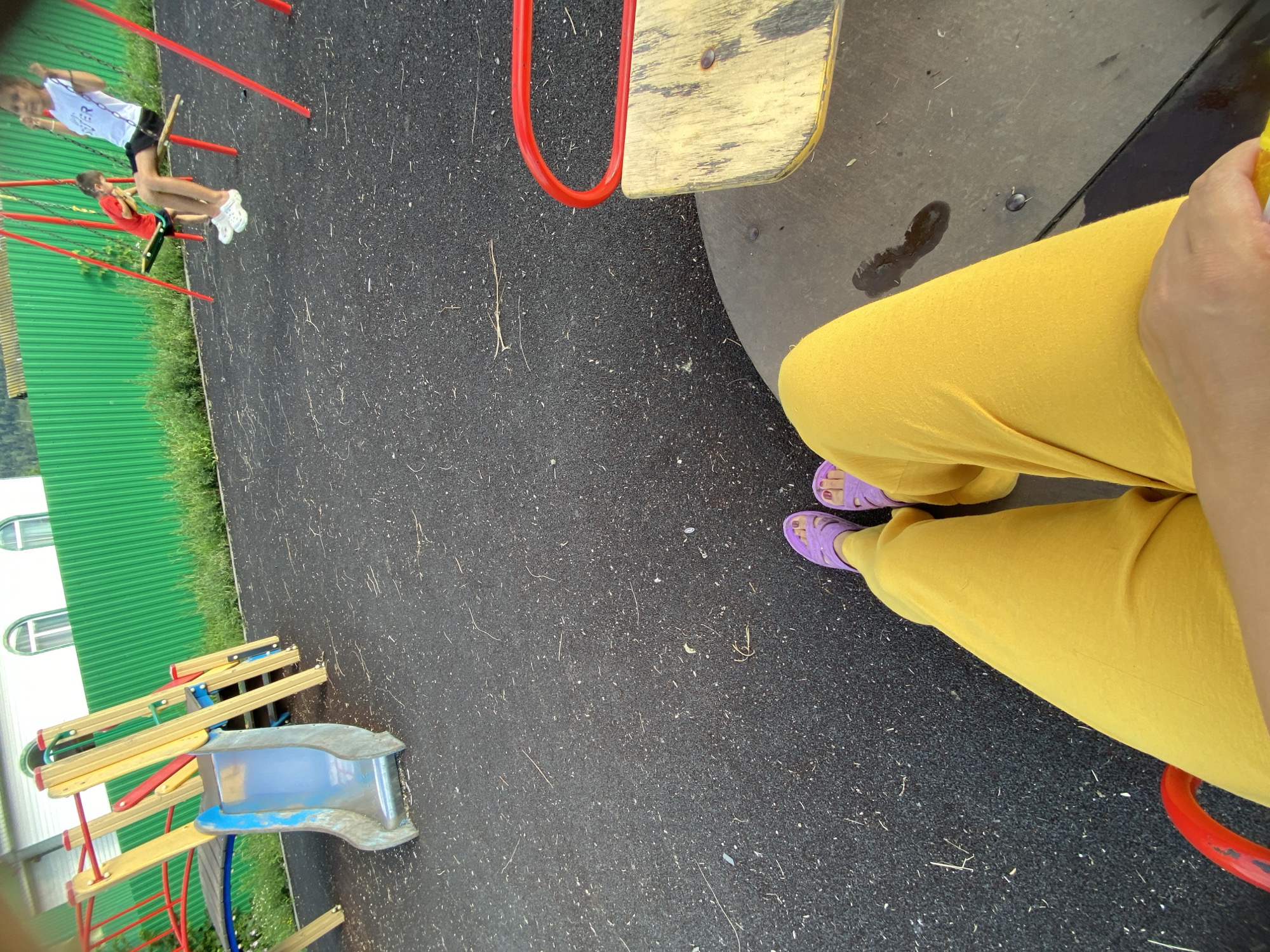 The image captures a scene at a children's playground in Maloarslangulovo, Russia. In the foreground, a person's legs clad in yellow pants and purple slides are visible, seated on a spinning ride. The ride itself has a yellow wooden seat and red metal bars. The ground is covered with a dark, rubberized surface. In the mid-ground, a blue plastic slide with yellow and red metal supports stands. Further back, two children are on swings. One child, wearing a red shirt, is on a swing with a green seat. Another child, in a white t-shirt, is on a swing with a wooden seat. A bright green corrugated metal fence runs along the background, with glimpses of vegetation and buildings behind it.