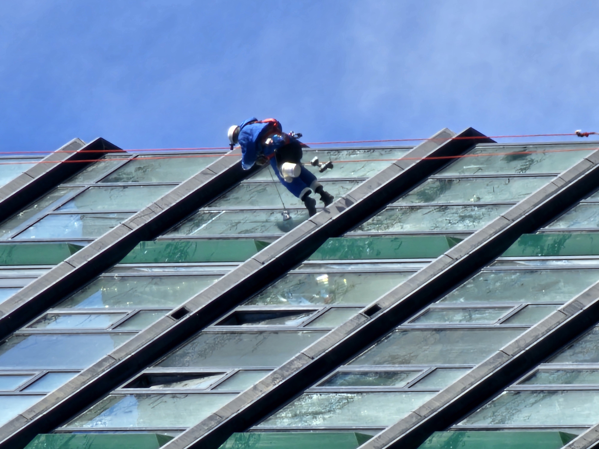 A window washer is rappelling down the side of a building with a geometric design. The building is located in the city of Moscow, Russia. The window washer is wearing a blue jumpsuit and a white helmet. He is using ropes and a harness to descend the building. The building has a series of angled glass panels with black frames. The sky is blue.