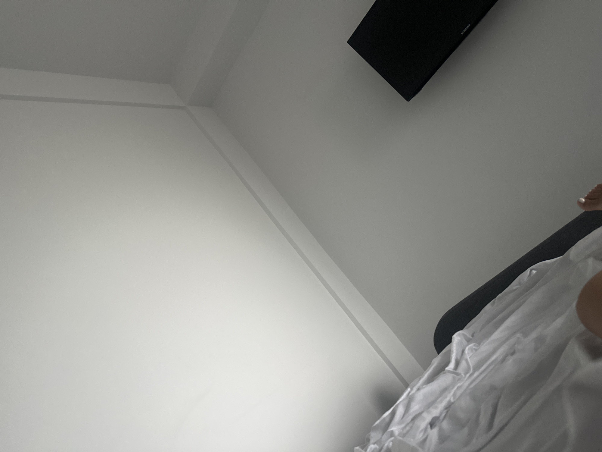 The image shows a corner of a room with white walls and a dark gray television mounted on the wall. There is also a white sheet or fabric draped over a dark object in the foreground. The lighting is dim, with a bright light source coming from the upper left corner of the image. There are no visible people in the image. The scene appears to be indoors, possibly a bedroom or living room. The location of Stațiunea Zoologică Marină Agigea, Romania, is not discernible from the visual information provided in the image.