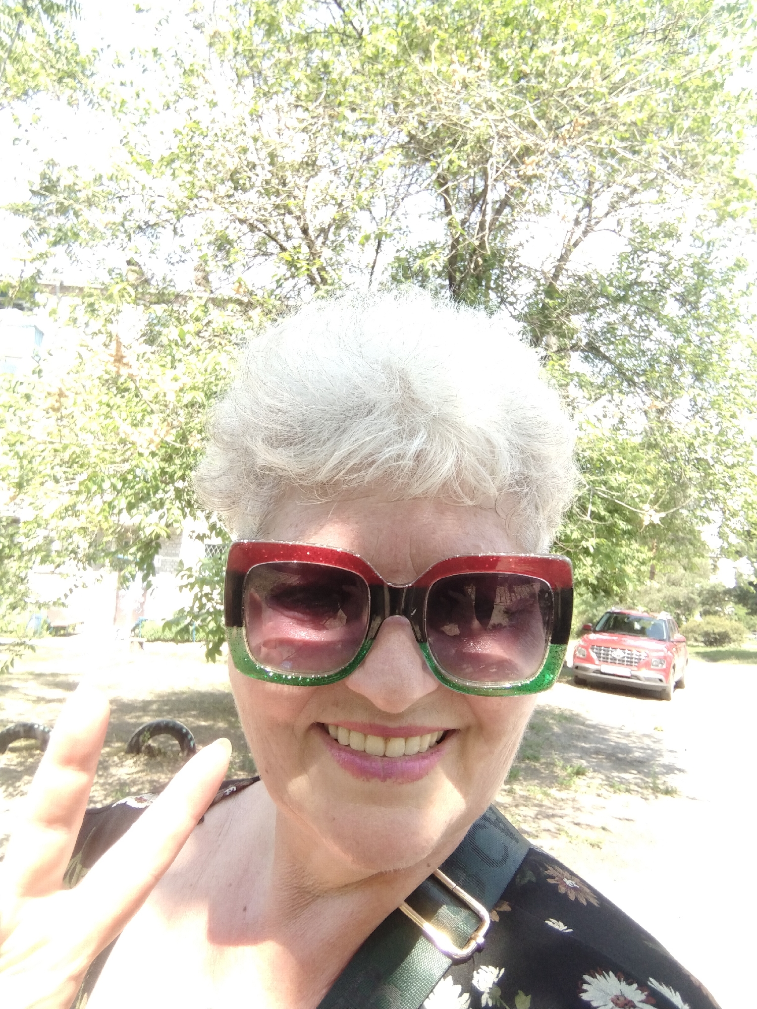 An adult woman with short, curly grey hair is captured in a close-up, upward-angled selfie, indicating self-photography. She is smiling broadly, revealing teeth, and wears oversized sunglasses with a red upper frame and a glittery green lower frame. Her left hand is raised, forming a "V" (peace) sign with her index and middle fingers. She is wearing a dark-colored garment, possibly a top or dress, adorned with a pattern of white or light-colored flowers. A green strap with a metallic buckle is visible across her right shoulder/chest area.
The background depicts an outdoor setting under bright daylight, with significant overexposure in parts of the sky. A large tree with green foliage and visible branches dominates the upper and right portions of the background. To the far left, a light-colored building is partially visible through the trees. In the mid-ground, to the right, a red compact SUV is parked facing the viewer on an unpaved, dirt or gravel path. On the ground to the left, two dark, worn tires are present, partially embedded in the earth. The scene is located in Horishni Plavni, Ukraine.