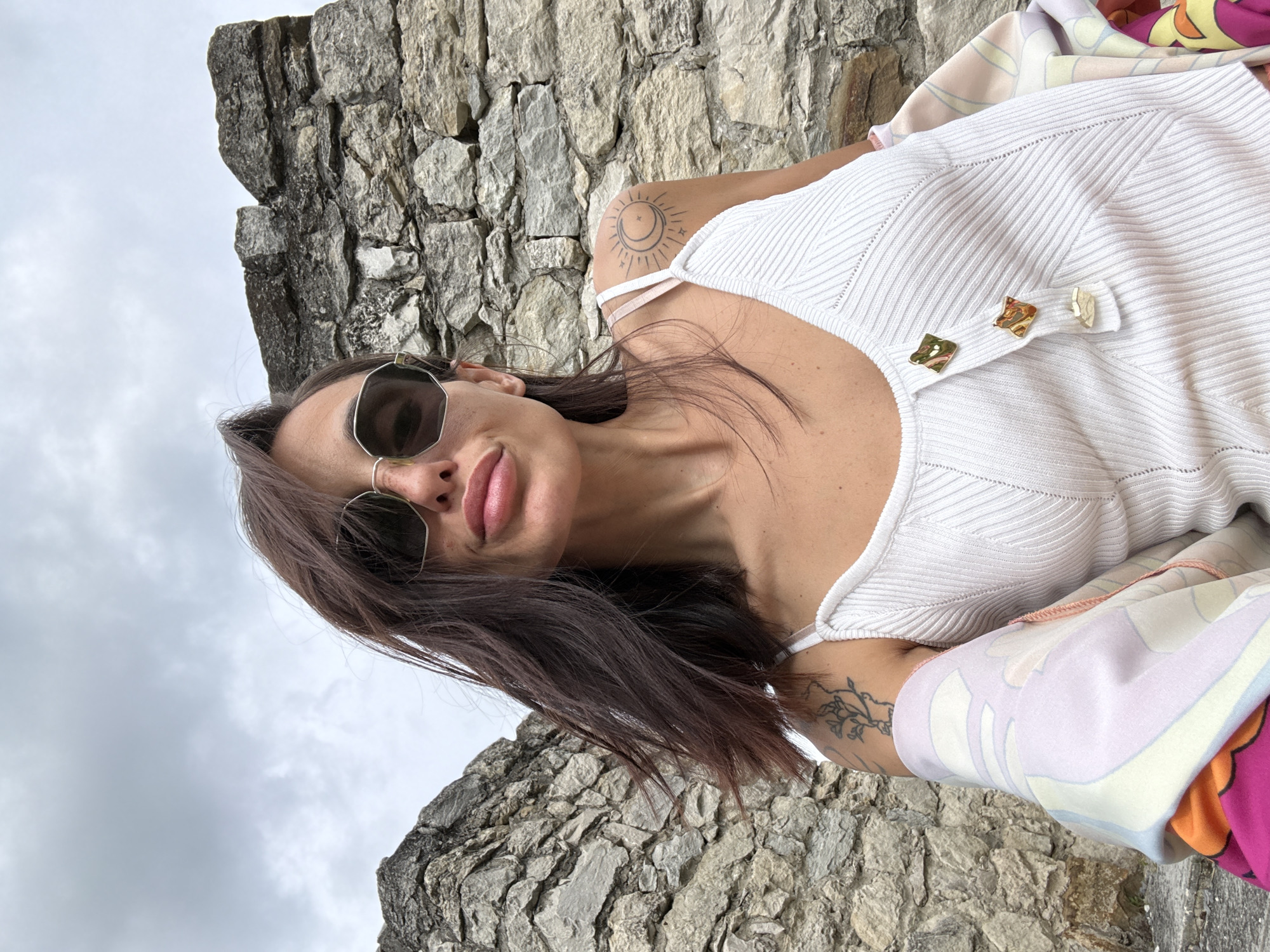 A woman with dark hair and sunglasses is posing for a selfie against a stone wall. She is wearing a white ribbed tank top with gold button details and a colorful patterned shawl draped over her shoulders. Tattoos are visible on her arms and shoulder. The background appears to be an outdoor setting with weathered stone structures. This location is likely within the Kaylaka Park in Pleven, Bulgaria, known for its ruins and natural scenery. The sky is overcast.