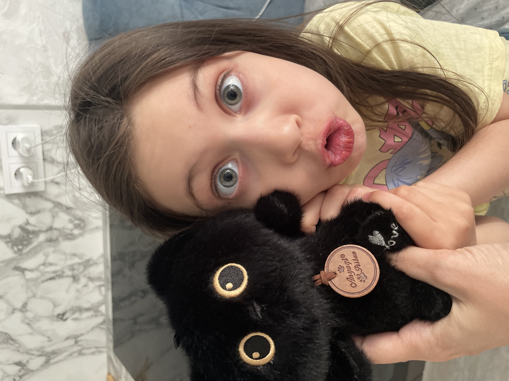 A photograph depicts a young girl indoors, likely in a domestic setting. The girl is holding a black stuffed animal, a toy that appears to resemble a black cat. The girl has long, dark hair and fair skin, with wide eyes and pursed lips. She is wearing a yellow t-shirt with a cartoon graphic. The stuffed animal features prominent, stylized eyes outlined in gold. A round tag with a brand name is visible, possibly "Anyaql Rubao," along with a paw print design. An electrical outlet is partially visible on the wall. The setting is undeterminable as Izhevsk, Russia from the given image.