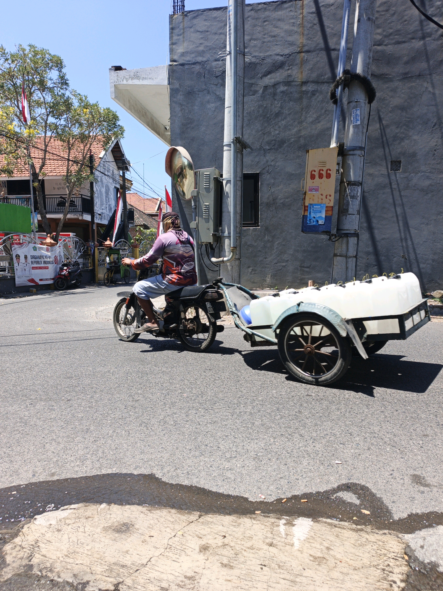 The image captures a daytime scene on a street in Ketegan, Indonesia, where a man is riding a motorcycle with a trailer attached. The motorcycle, a dark-colored model, is being ridden by a person wearing a patterned long-sleeved shirt and light-colored shorts, with a bandana covering their head. The trailer, a light-colored, rectangular unit with a sloped front, is laden with multiple white plastic containers, each with yellow caps. The trailer has two wheels, one of which is prominent with a spoked design. The motorcycle and trailer are moving on an asphalt road.
The environment includes buildings with grey and white facades. One building has a textured grey wall with a large white overhang. There are trees with green foliage and some yellow flowers visible, along with red and white Indonesian flags. A utility pole with various attachments and signs, including one with red "666" numerals, stands near the road. The sky is clear and blue. The road surface shows some wet patches and discarded cigarette butts near the curb. In the background, there are banners and other vehicles, including a parked red motorcycle and a bicycle, suggesting a moderately busy urban setting. The lighting indicates it is a sunny day, casting shadows on the road.