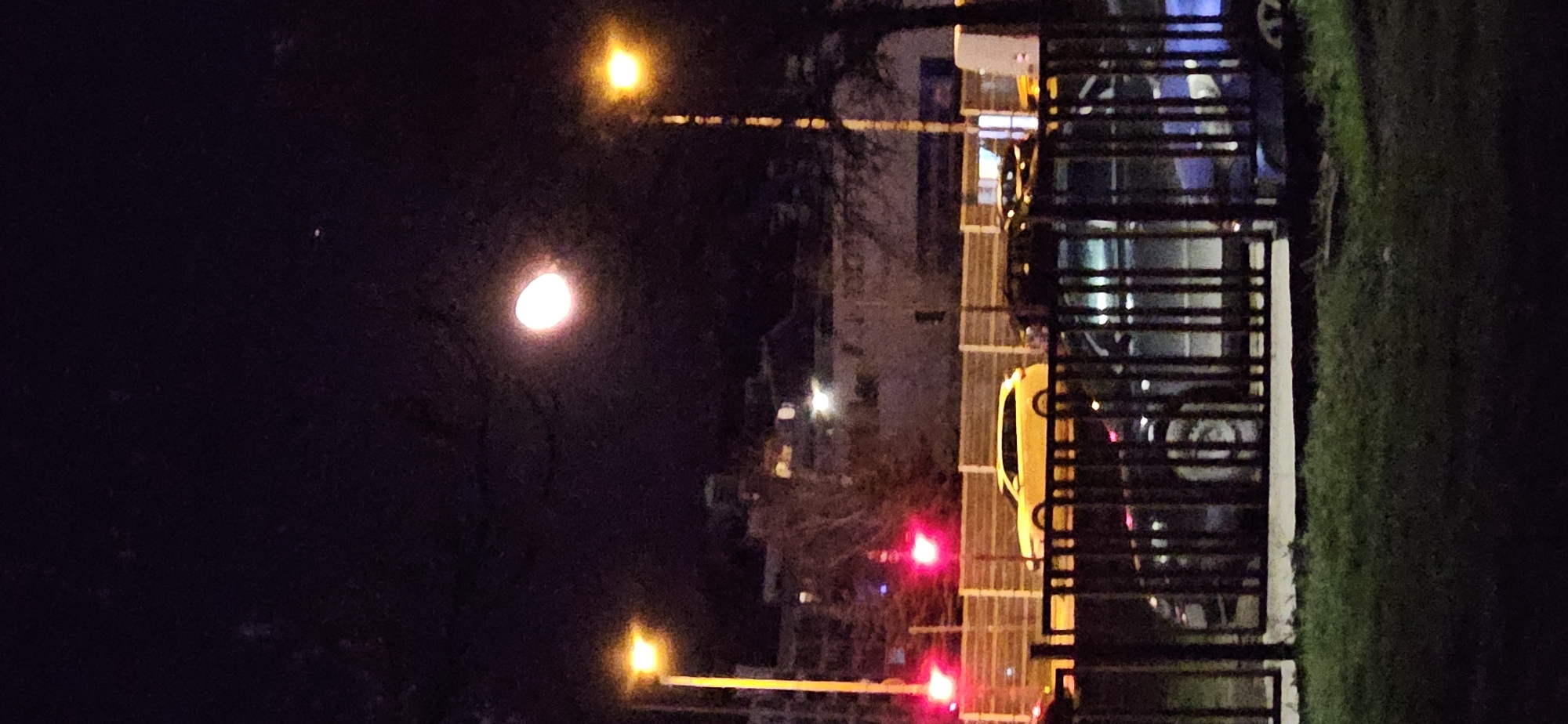 The image depicts a nighttime scene in an urban environment, likely Sofia, Bulgaria. Dominating the left portion of the frame is a dark sky with a bright moon and a nearby street lamp. In the central and right areas, a fence runs vertically. Through the fence, there are parked cars including a yellow car that is closest to the fence. Street lights and traffic lights illuminate the scene, adding to the urban ambiance. The ground on the right side of the fence is covered in grass.