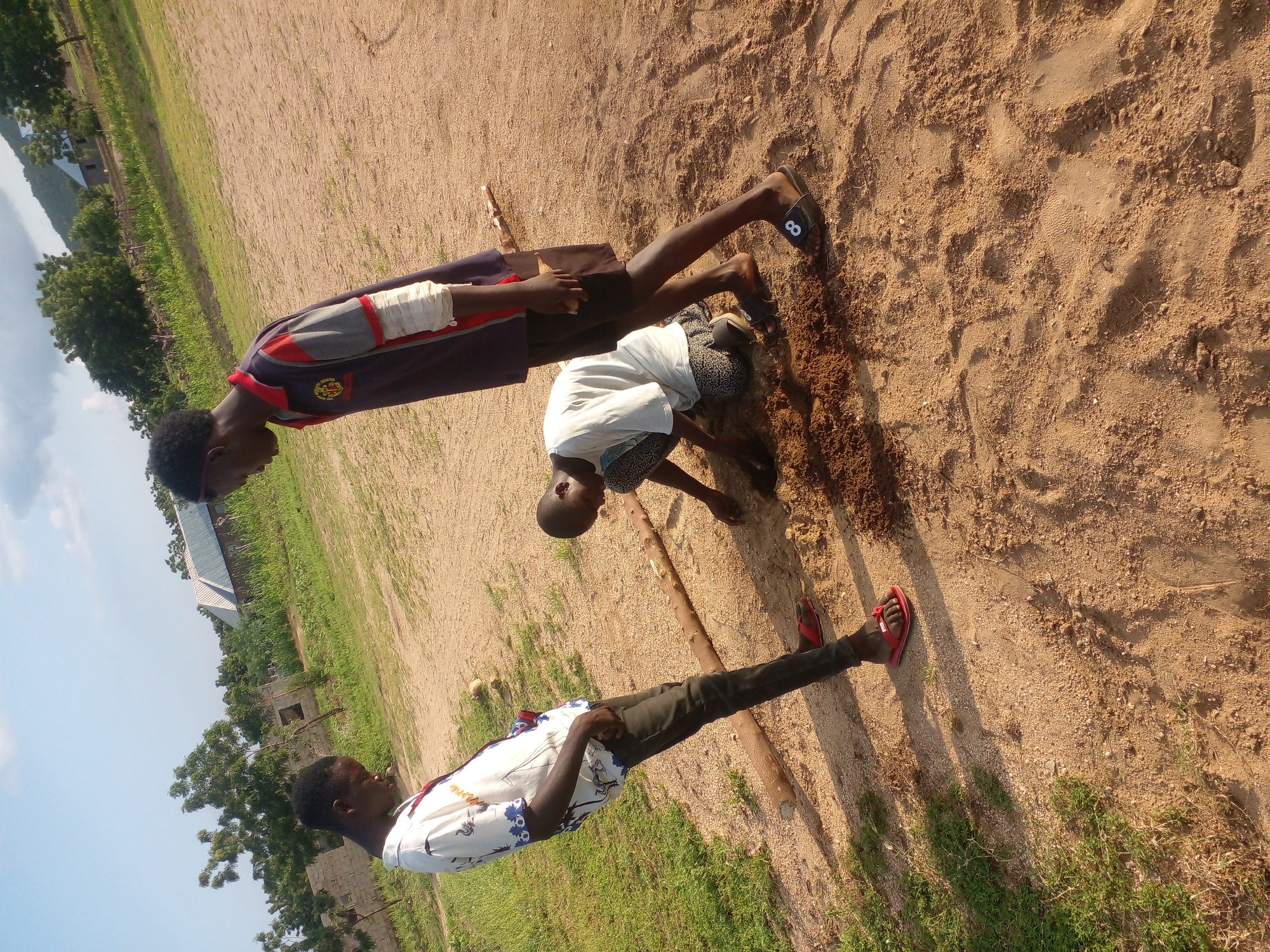 The image depicts three young boys in an outdoor, rural setting. The ground is composed of dry, sandy soil with patches of sparse green grass. In the background, there are some partially constructed buildings, trees, and a distant hill under a partly cloudy sky. The primary focus is on the three boys. One boy, wearing a white patterned shirt and dark pants, stands on the left, looking down. Another boy, in a dark polo shirt with red and grey accents and dark shorts, stands on the right, also looking down. His left arm is bandaged. The third boy is crouched in the center, actively digging or manipulating the soil with his hands. A long, light-colored wooden pole lies horizontally on the ground between the standing boys and the crouching boy.