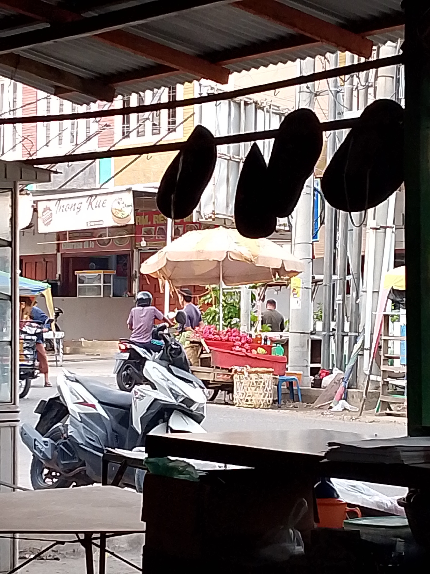 The image captures a street scene in Lhokseumawe, Indonesia, with a focus on a food stall and its surroundings. In the foreground, a portion of a wooden table and a dark object, possibly a bag, are visible, suggesting an indoor vantage point looking outwards. Outside, a white and grey scooter is parked prominently. Further back, a street unfolds with a few individuals and vehicles.
On the street, a person wearing a purple shirt and a helmet is seated on a scooter. To their right, another person in a dark shirt stands near a stall covered by a light-colored umbrella. This stall displays a red container filled with what appears to be red fruits, possibly apples or pomegranates, and a woven basket. Behind the stall, a small cart is visible.
Further down the street, a woman in a blue jacket and brown pants is standing next to a vehicle with a blue awning. Another scooter is parked nearby. The background reveals buildings with a striped facade, suggesting a commercial area. Overhead, a corrugated metal roof structure, supported by beams, extends across the scene. Several dark, oval-shaped objects are hanging from a horizontal bar, silhouetted against the sky. The overall impression is of a bustling, everyday scene at a street-side marketplace.