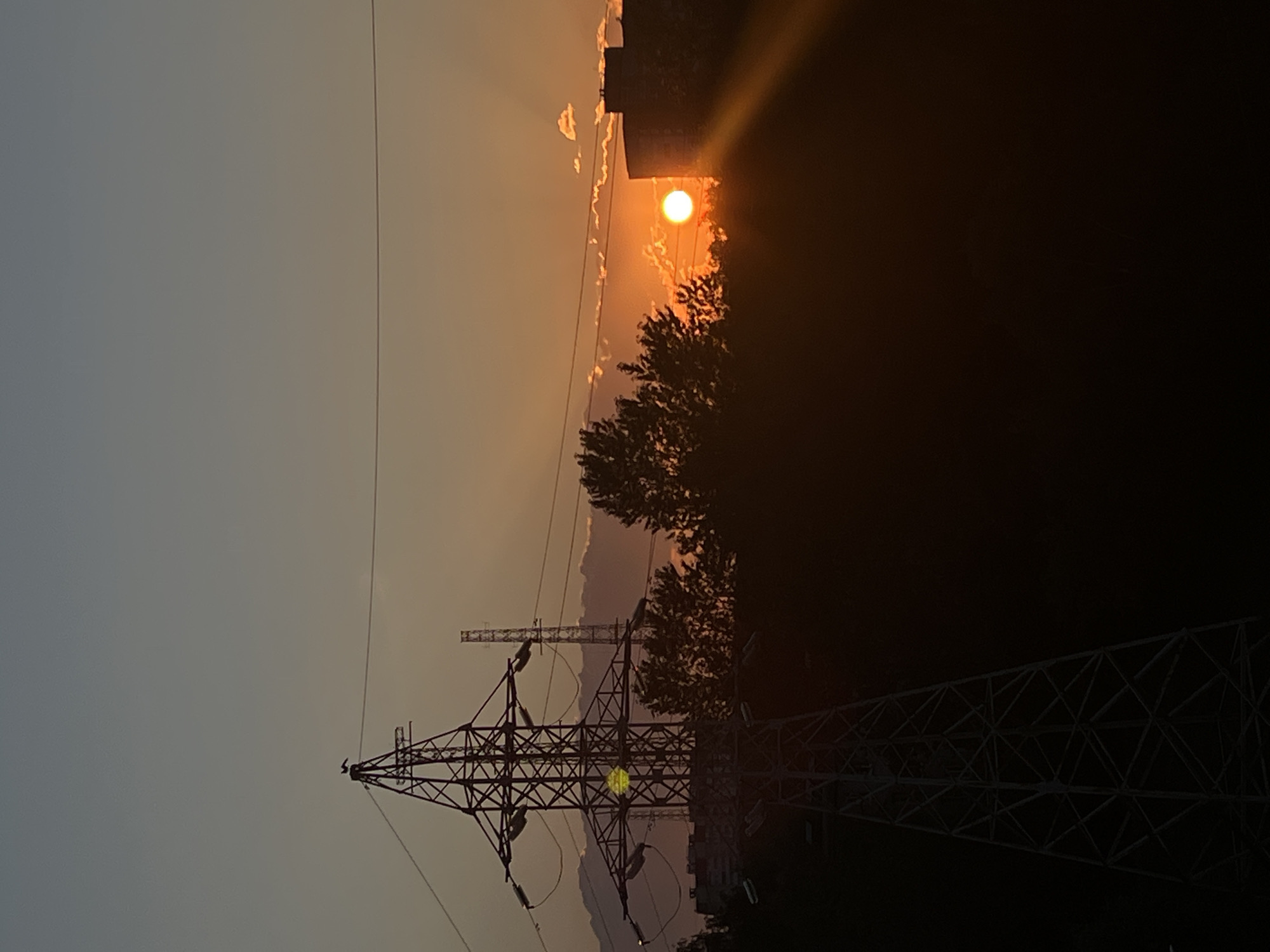 The image depicts a sunset scene in Saint Petersburg, Russia. The sun, a bright orange orb, is setting behind trees and a building, casting dramatic rays of light through the clouds. In the foreground, a large electrical pylon with intricate metal lattice work is silhouetted against the darkening sky. Another, smaller pylon is visible in the distance. The overall mood is one of tranquility and the end of the day. The sky transitions from a pale grey on the left to a warm orange and yellow gradient on the right, indicating the sun's position near the horizon. The dark silhouettes of trees and buildings suggest a semi-urban or industrial landscape. There is no visible text in the image.