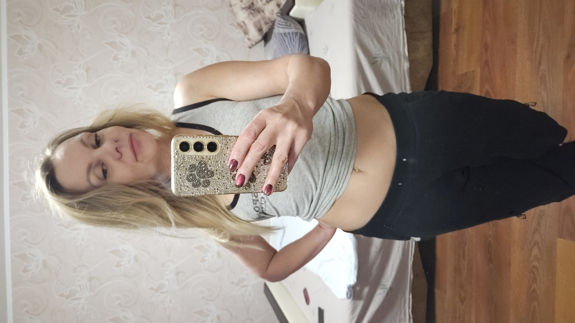 A woman is taking a selfie in front of a mirror. She is wearing a gray tank top and black pants. She is holding a smartphone with a decorative case. The background includes a bed, a wall with patterned wallpaper, and a wooden floor. The location is likely indoors and residential. There is no indication of the city of Sevastopol, Ukraine.