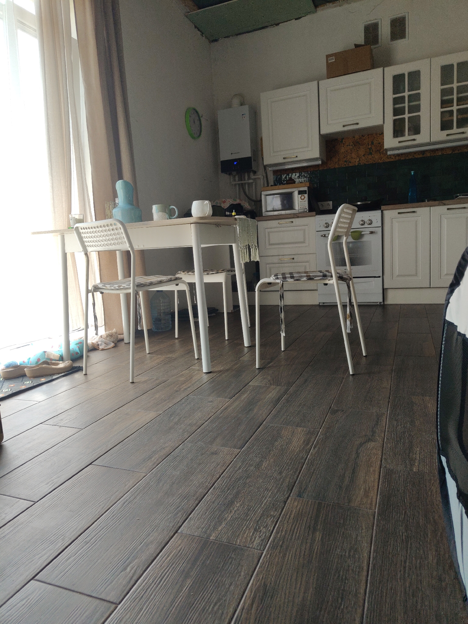 The image shows a kitchen with a dining table and chairs. A white dining table is set with two chairs, one with a checkered cushion. There are mugs and a pitcher on the table. A window with sheer curtains is to the left. The kitchen cabinets are white, and a white oven is visible. A microwave sits on the counter. The floor is covered in dark wood-look tiles. The image is a low-angle shot, looking across the floor towards the kitchen area. The location is stated to be Yuzhny, Russia.