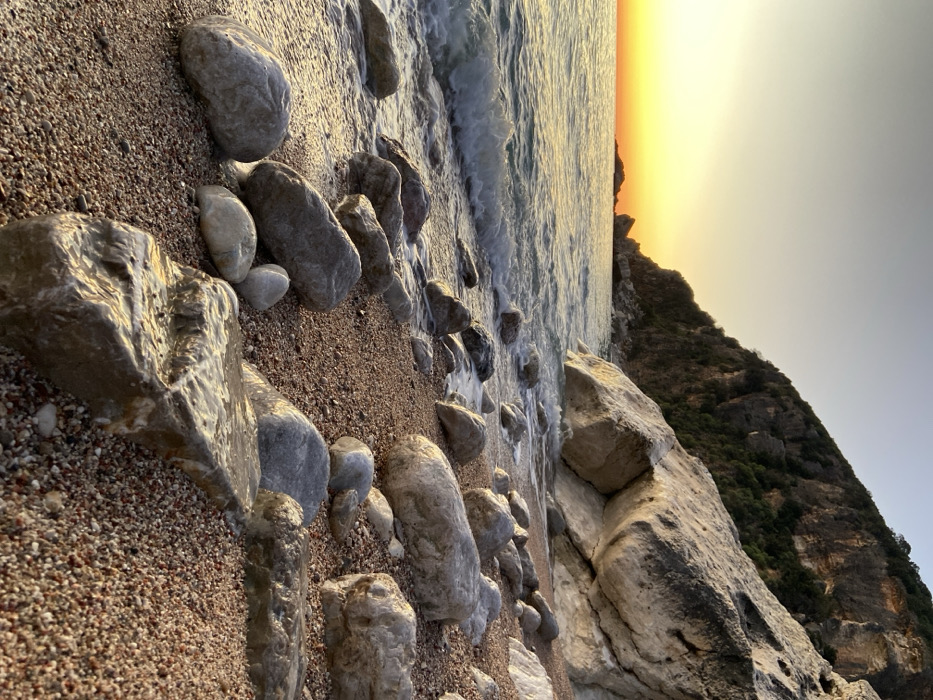 The image displays a coastal landscape during golden hour, likely sunrise or sunset, located in Unknown, Unknown. The scene is dominated by a pebbled beach in the foreground, composed of reddish-brown gravel mixed with numerous smooth, irregularly shaped rocks ranging from grey to off-white. Several of these rocks near the waterline are visibly wet, reflecting the light. Small waves with white foam are breaking and receding along the shore on the left side. The water body, appearing as a sea or ocean, stretches towards the horizon, reflecting a strong orange and yellow glow from the low sun, which creates a vibrant gradient in the sky transitioning to a lighter blue-grey overhead. In the distant water, small rocky outcrops are visible. To the right, a large, steep cliff face rises dramatically from the beach, characterized by lighter-colored rock formations and patches of dark green vegetation on its upper sections. No individuals or man-made structures are present.