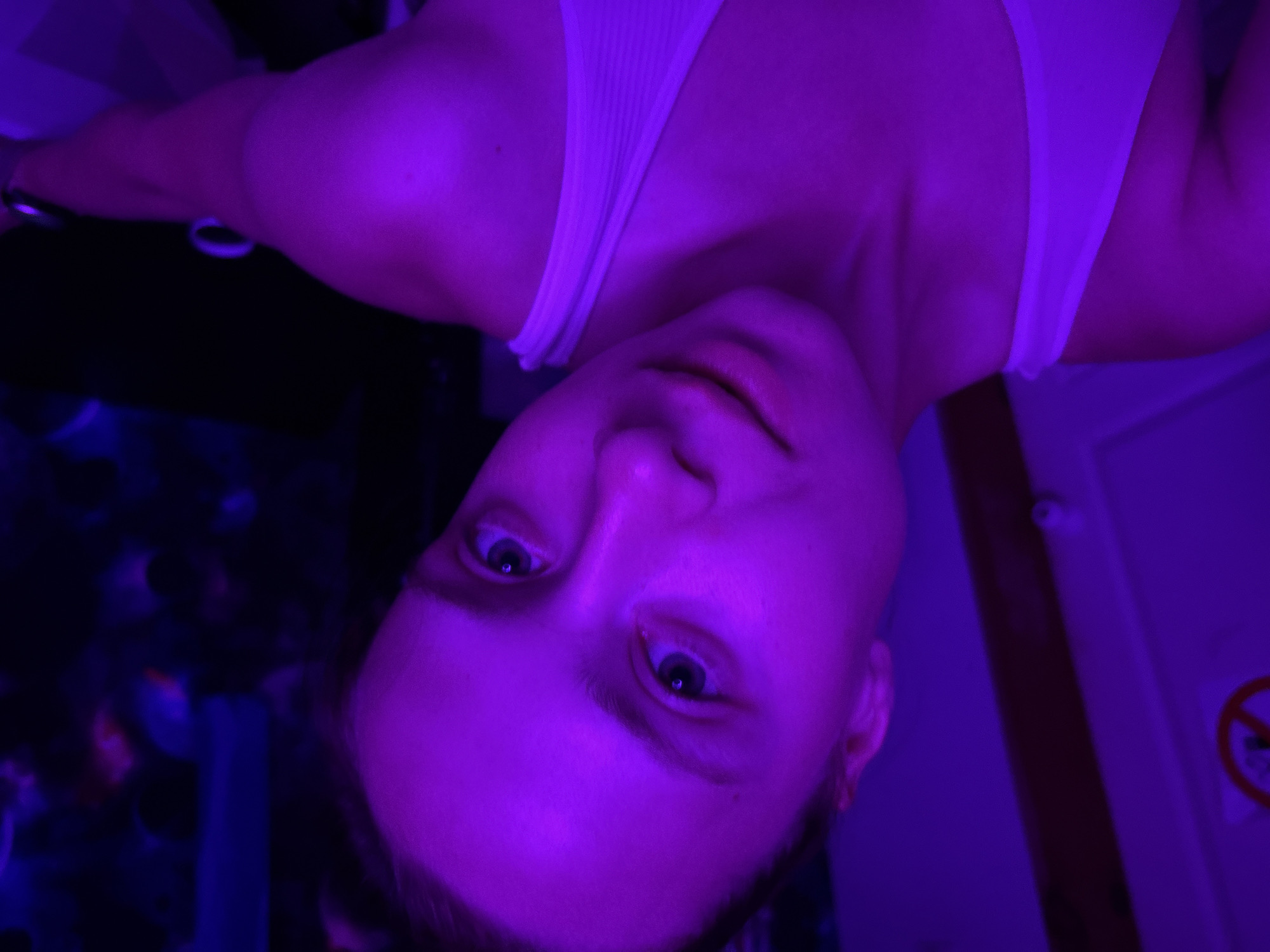 A medium shot captures a person with pale skin, viewed from below and at an angle, set against a backdrop of what appears to be a room interior. The individual is lit in purple lighting, which casts a strong hue over their face and upper body. They are wearing a white tank top. The camera angle makes the person appear upside down. In the periphery, there is a partially visible area with multicolored, blurred shapes resembling a patterned surface and part of a door with a no smoking sign on it. The location is Прикамский, Russia.