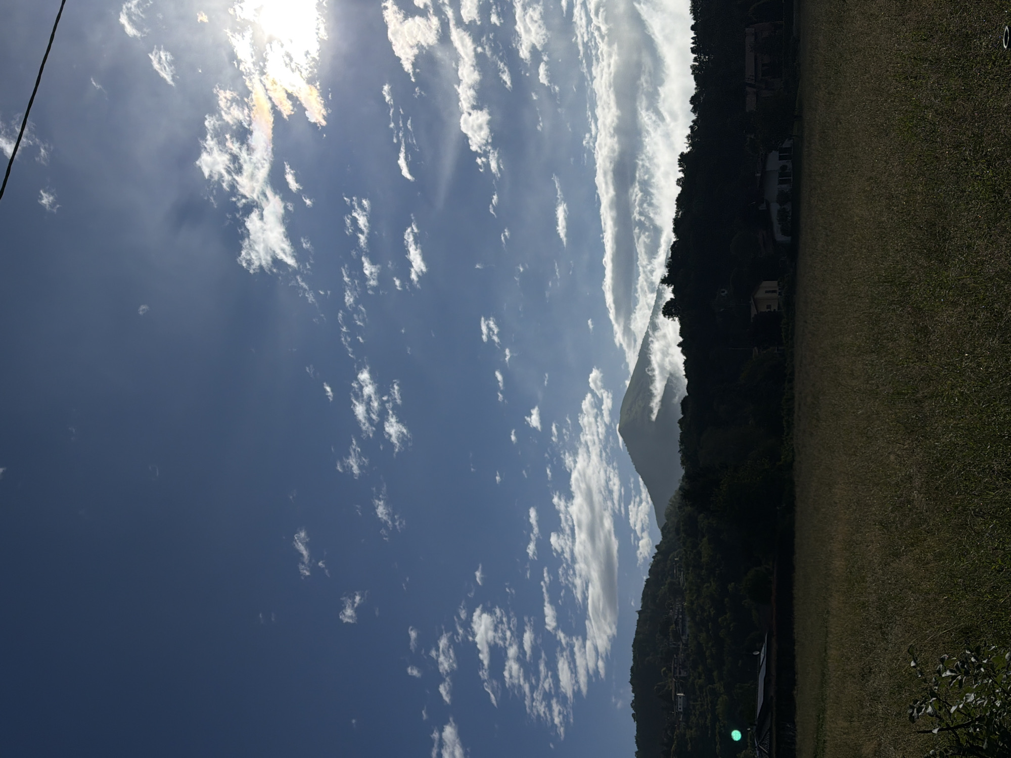 The image depicts a daytime landscape near Penzano, Italy. A field of grass occupies the right side of the image. Adjacent to the field is a densely forested hillside with scattered houses visible among the trees. A mountain peak rises above the trees, partially obscured by clouds. The sky is blue with numerous white clouds. The sun is visible near the top of the image, causing some glare. A power line is present in the upper left corner of the image.