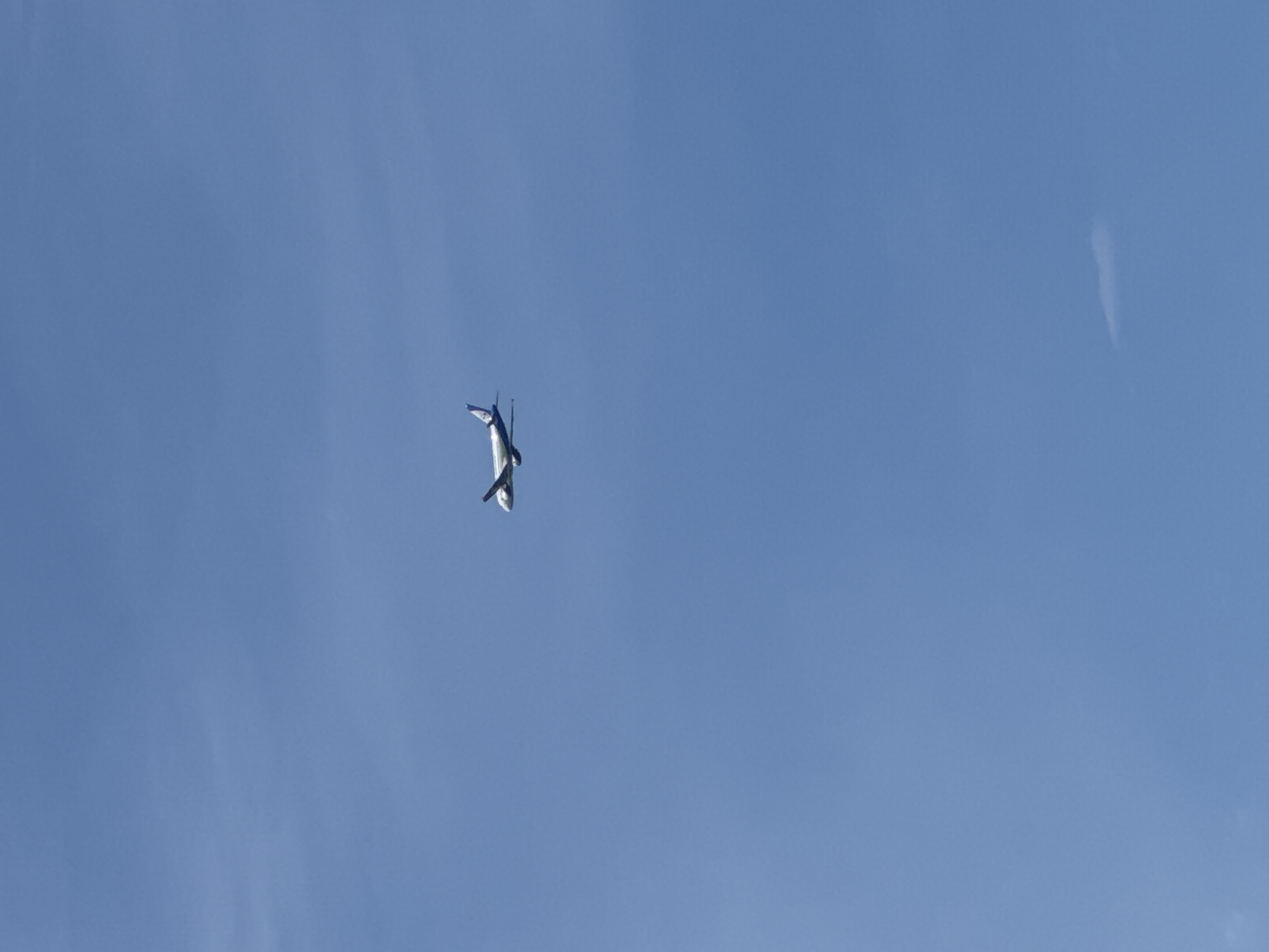 A low-angle, wide shot depicts a clear blue sky with a small aircraft in the left center. The aircraft appears to be white with blue markings on its tail. The sky is almost cloudless, with a few faint wisps of clouds. No people are visible. The location is unknown and cannot be determined from this image.