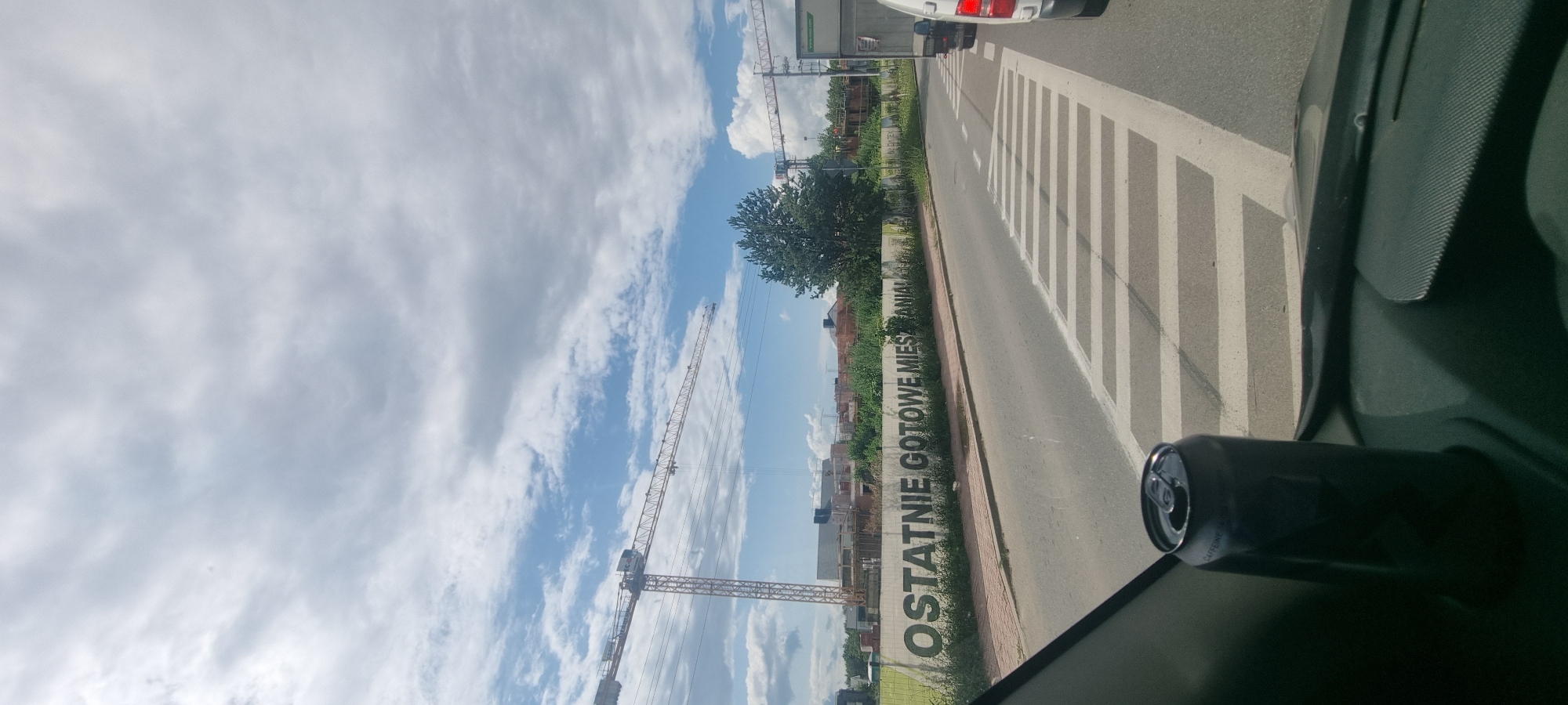 In Otrębusy, Poland, a concrete wall along a road displays large, dark lettering that reads "OSTATNIE GOTOWE MIESZANIA". The wall is situated behind a sidewalk and some vegetation. The scene appears to be a construction area, with cranes visible in the background, suggesting ongoing development. A road with white crosswalk markings is in the foreground, with traffic lanes visible. A dark-colored beverage can rests on the interior dashboard of a vehicle, partially obscuring the view of the road ahead. The sky is partly cloudy, with a mix of blue sky and white clouds.