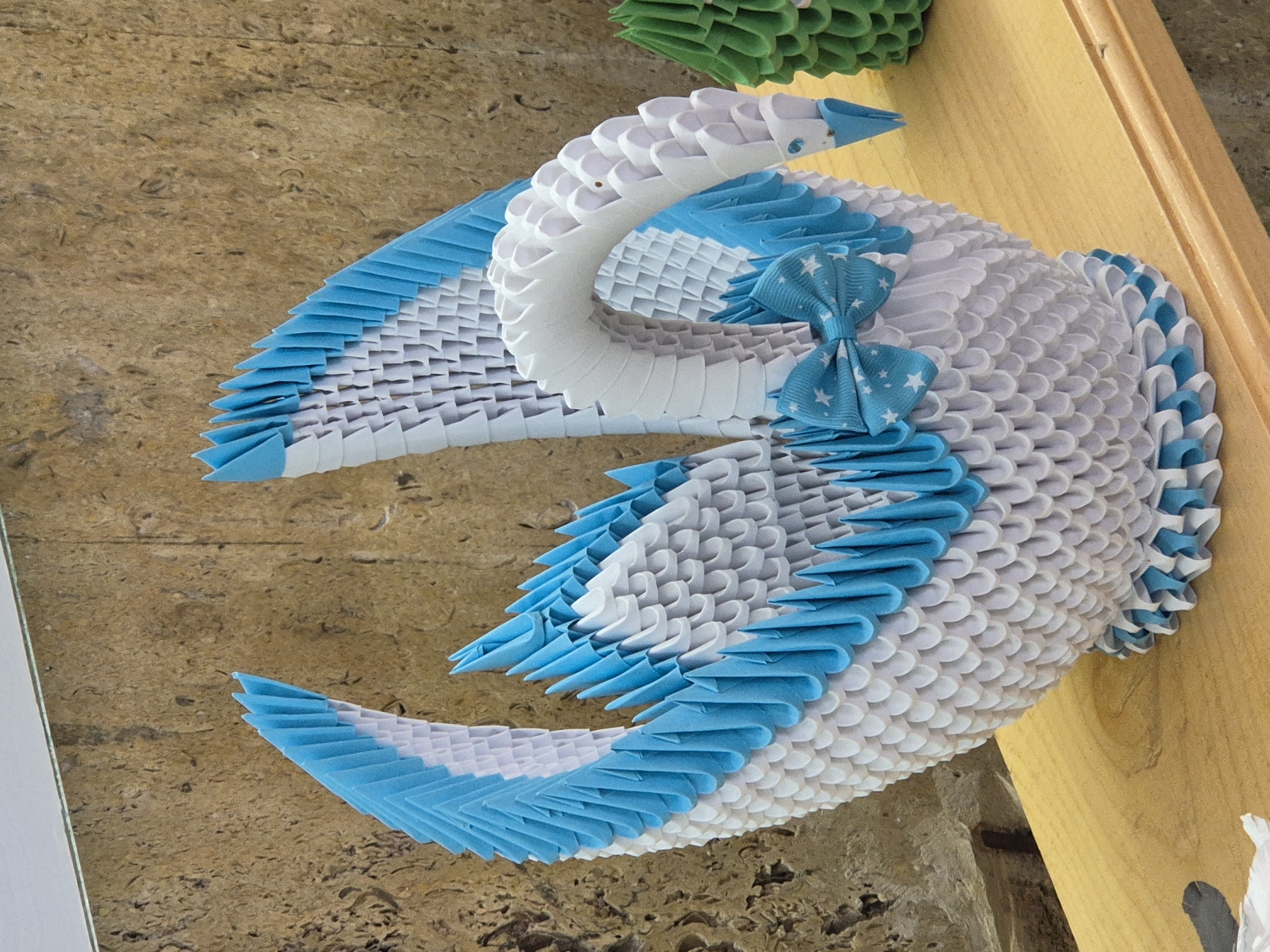 The image shows a 3D origami swan, primarily white with blue accents along the edges of its wings and tail, and a blue bow with white stars around its neck. The swan is placed on a surface composed of wooden planks and a wall with a textured beige surface. A small portion of another origami structure, green in color, is visible in the upper part of the frame. The precise location within Srebarna, Bulgaria, cannot be ascertained from this image.