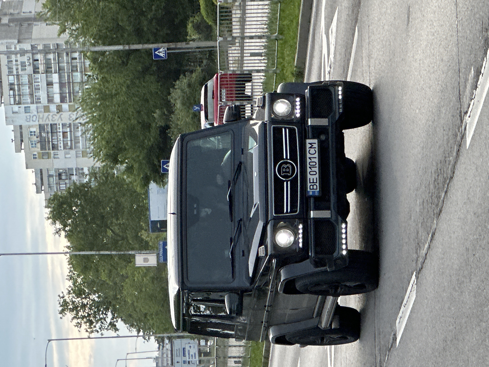 A black Brabus G-Wagon with license plate "BE 0101 CM" is centered on a light gray asphalt road. It has LED lights on its front and its headlights are on. The background includes trees and a tall residential building with the Cyrillic writing "З УЗУНОВ." The scene is located in Varna, Bulgaria, as indicated by the Cyrillic inscription and the license plate code. A pedestrian crossing sign is visible.