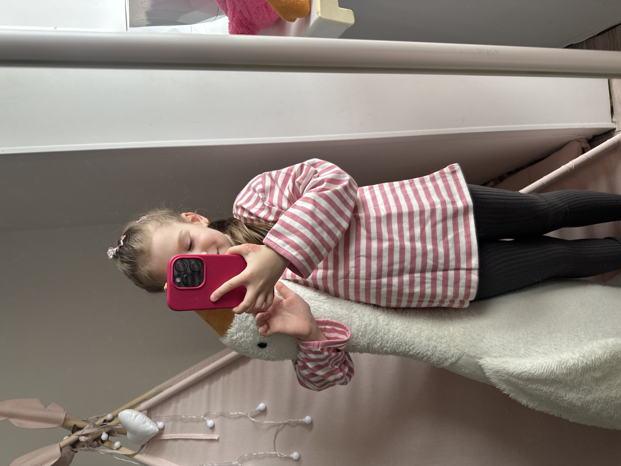 A young girl in a pink and white striped top and black leggings is sitting on a white stuffed goose within a pink teepee-like structure. She is holding a pink smartphone, presumably taking a selfie. The background includes a white shelf or bunk bed frame. The location is likely a home interior, possibly in the city of Chaikovsky, Russia.
