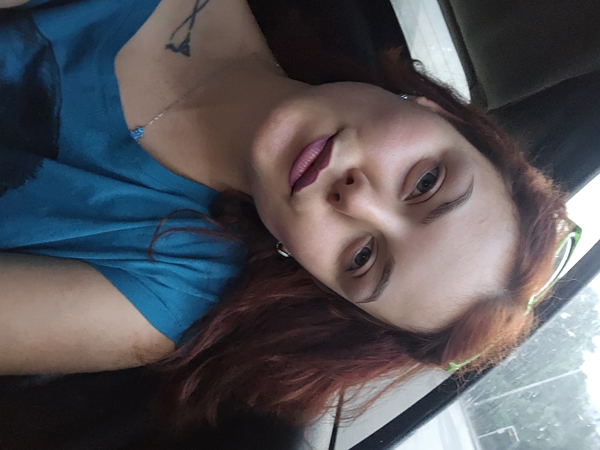 A young woman with red hair, wearing a blue t-shirt, and makeup, is photographed inside a vehicle. The woman is in the foreground, facing the camera, with her head and shoulders visible. She is wearing a blue t-shirt, a silver necklace with a blue pendant, and earrings. She has a tattoo on her left shoulder. Her hair is long and wavy. She is wearing lipstick and has her eyebrows done. The background shows the interior of a vehicle, with a window showing a blurred outdoor scene. The photo appears to be a selfie taken by the woman. The location is unknown.