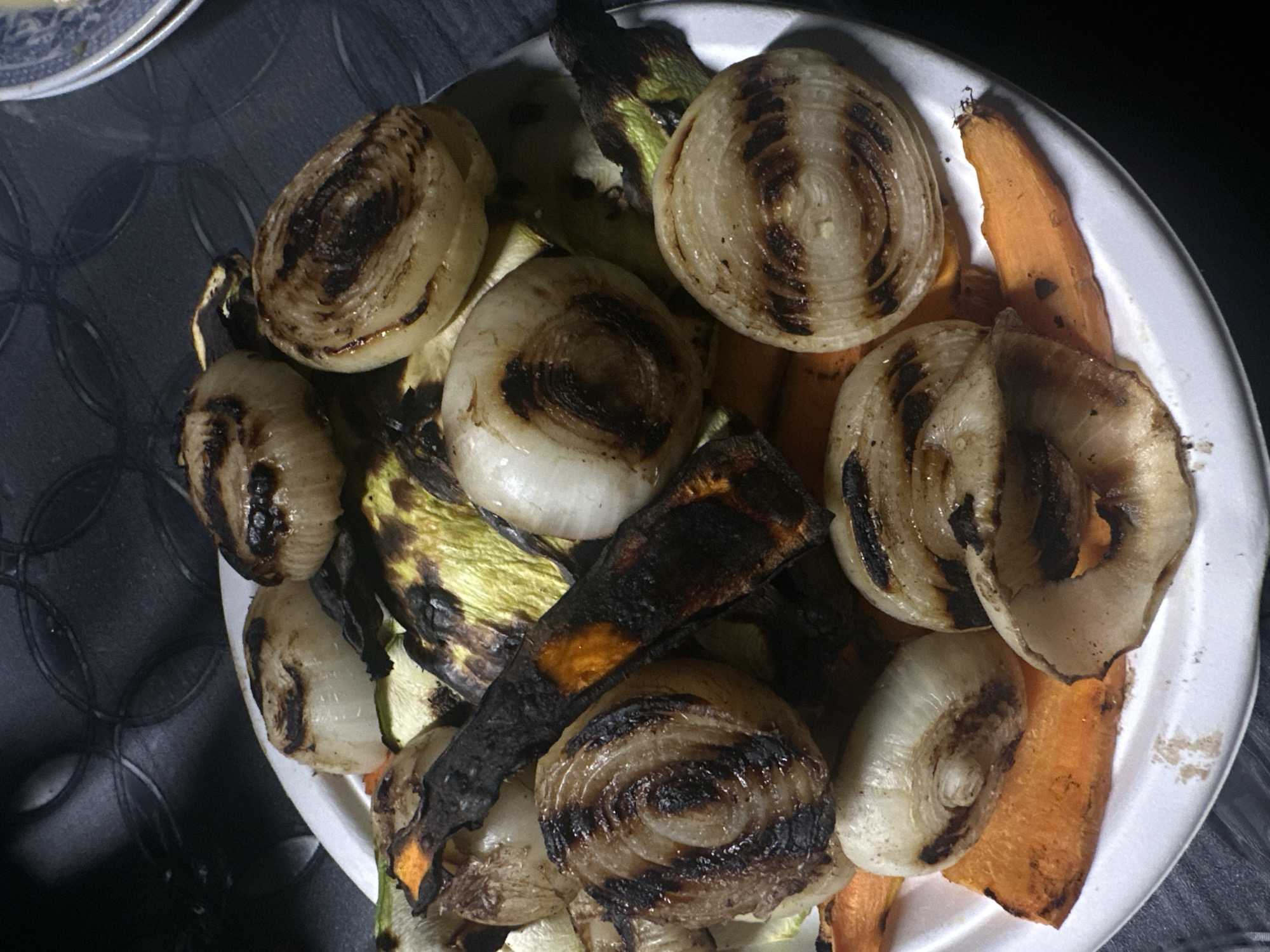 A white, possibly disposable, plate is densely arranged with an assortment of grilled vegetables, located in Svetovrachene, Bulgaria. The plate contains numerous small, whole or halved onions, displaying distinct dark brown to black charring and grill marks, often in concentric patterns, against their soft, pale cooked surfaces. Bright orange carrot slices or sticks are interspersed throughout, some with significant dark charring. Pieces of green and yellow squash or zucchini are also present, marked with dark grill lines. A visibly charred, long piece of carrot appears predominantly black with a bright orange interior. At least one light brown, grilled mushroom cap is observable. The plate rests on a dark, textured surface, possibly a tablecloth, which features a subtle pattern of interwoven circles. The scene is illuminated by an unseen light source, casting soft shadows and highlighting the cooked textures of the vegetables. A small portion of a patterned blue and white plate is visible in the upper right background.