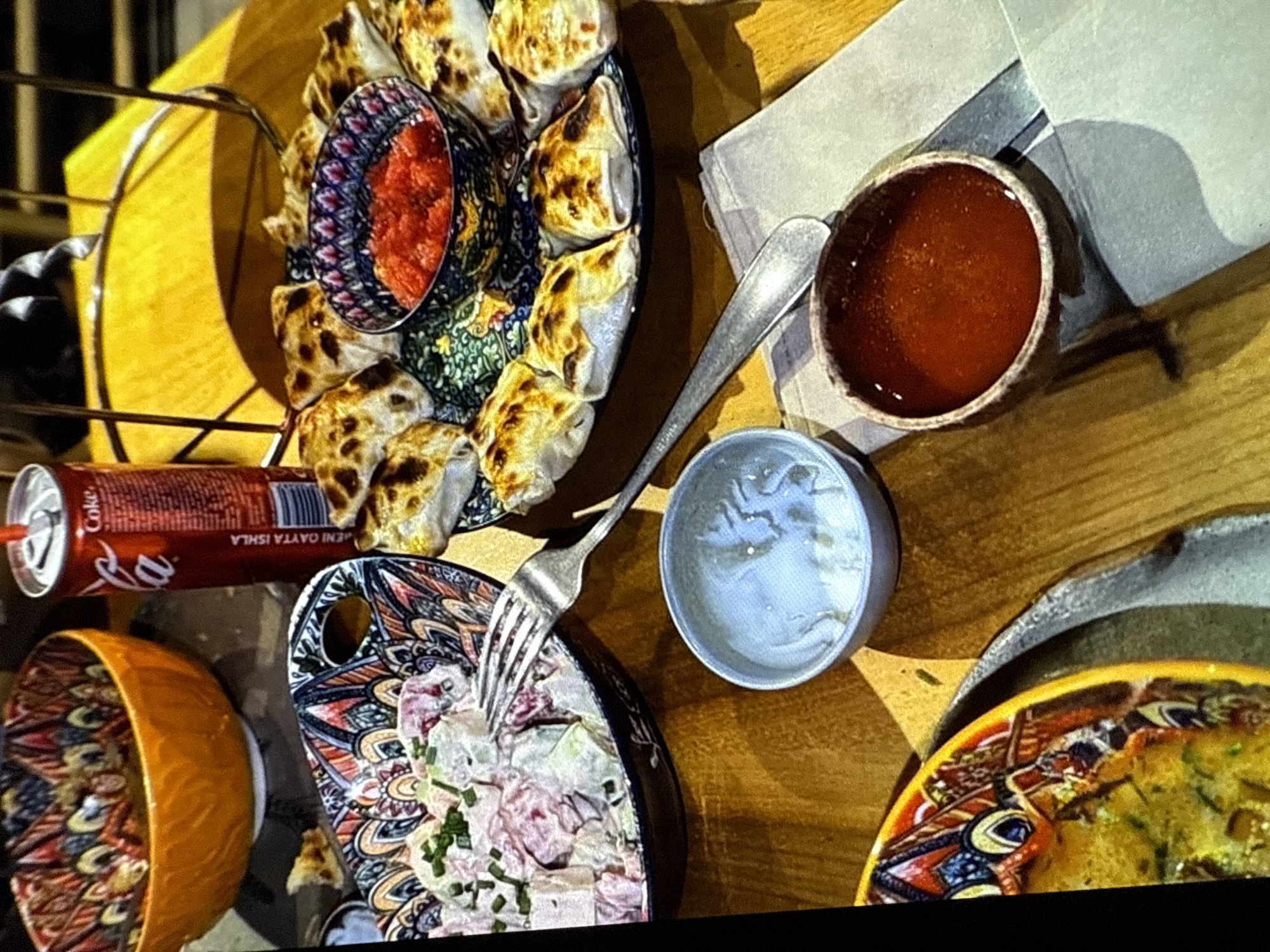 A close-up, overhead shot captures a culinary spread on a wooden table. In the upper left, a vibrant orange bowl, intricately decorated with a colorful mosaic pattern, is filled with food. To its right, a red Coca-Cola can stands with a straw. A large platter, also adorned with a similar mosaic design, features a circular arrangement of golden-brown, crescent-shaped pastries surrounding a small, dark blue bowl filled with a bright red sauce. In the foreground, a fork rests on the table, with its tines submerged in a bowl of light-colored salad topped with chives. Beside it, a small, grey, ceramic bowl contains a small amount of white residue. Further to the right, a small, rustic brown bowl holds a dark red, viscous liquid, possibly a sauce or dressing, resting on a folded light grey napkin. In the lower left corner, a portion of another vibrantly decorated yellow bowl is visible, containing a greenish-yellow dish. The lighting suggests an indoor setting, possibly a restaurant.
