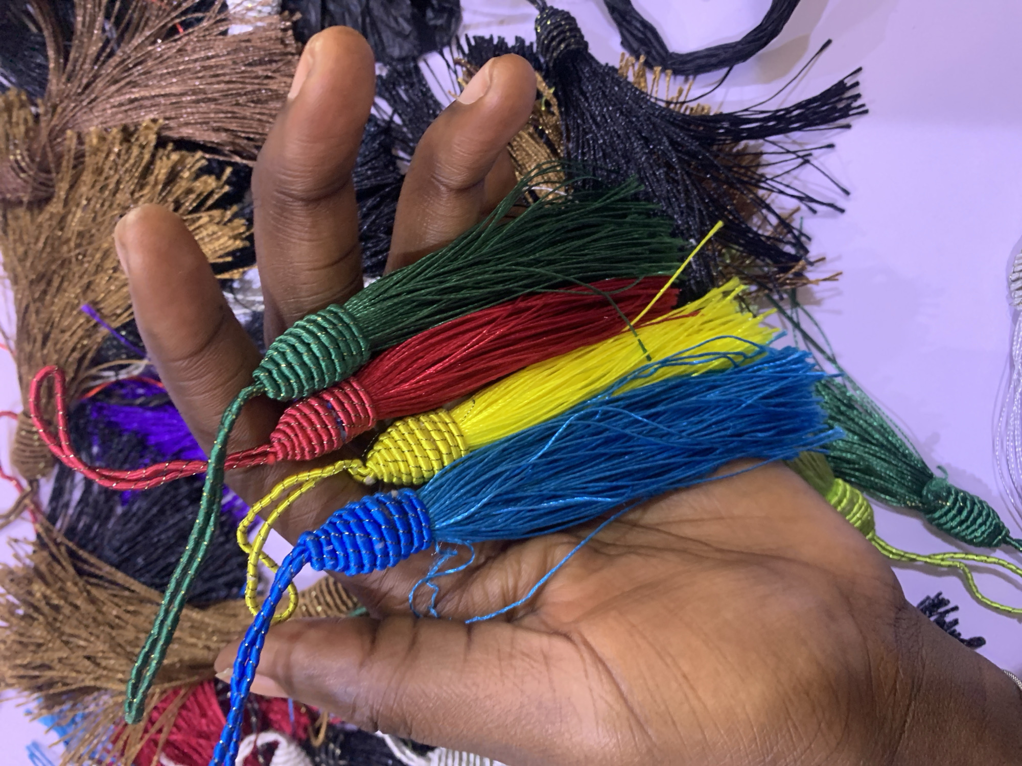 A person's hand is holding a collection of multi-colored tassels, primarily blue, yellow, red, and green. The tassels have a braided top and loose threads at the bottom. The background appears to be a pile of similar tassels in various colors, including brown, black, and purple. The image is taken from a close-up perspective, focusing on the hand and the tassels it holds. There is no visible indication of a specific location in Gura Top, Nigeria.