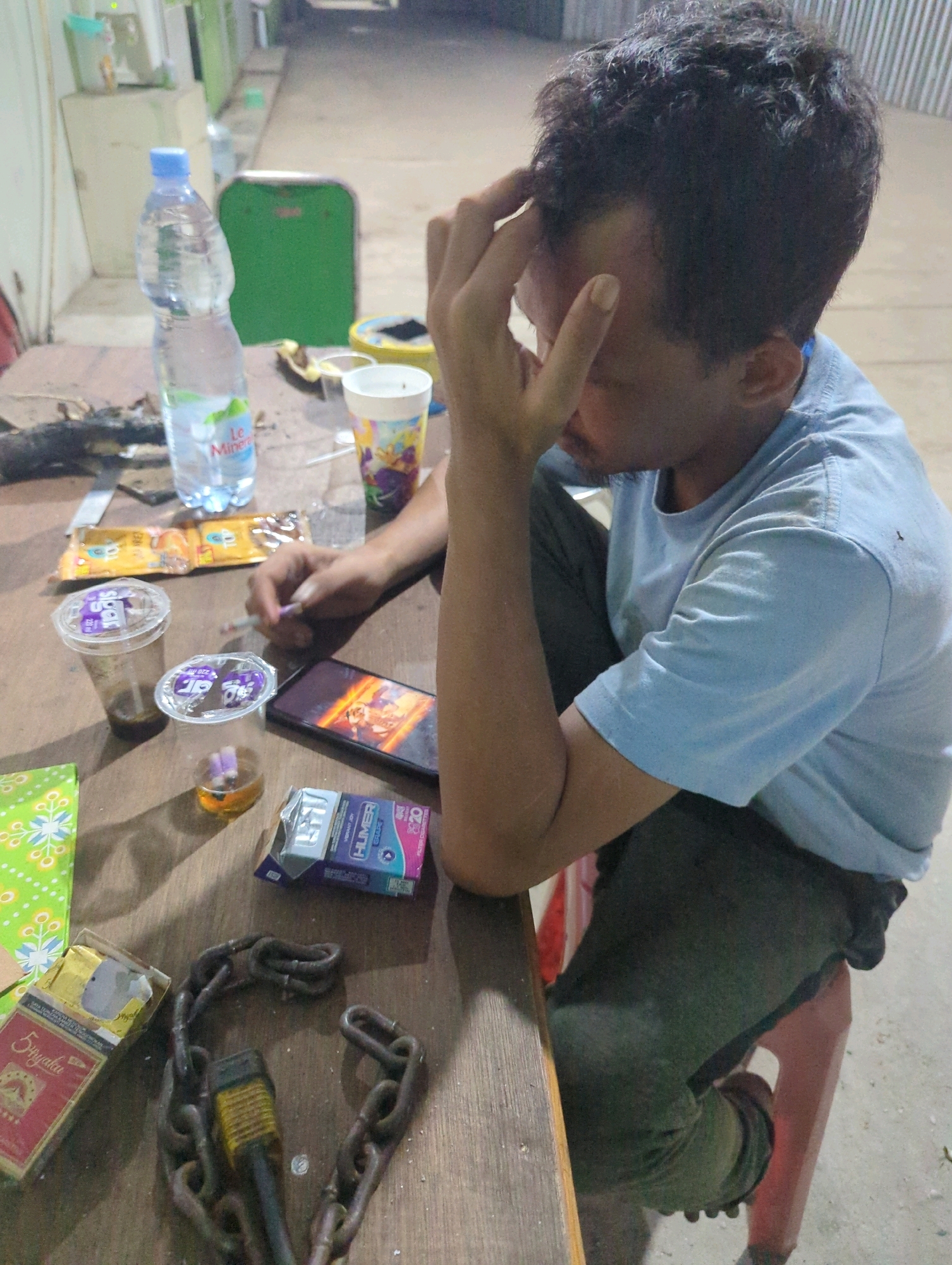 In Batu Belaman, Indonesia, a man with dark, curly hair sits at a wooden table, his head bowed and hand to his forehead, suggesting distress or fatigue. He wears a light blue t-shirt and dark pants. The table is cluttered with various items: a partially opened pack of "555" cigarettes, a pack of "Hummer" cigarettes, two plastic cups containing drinks and what appear to be cigarette butts, a smartphone displaying a vibrant image, a bottle of "Le Minerale" water, and two sections of a heavy chain. A green, somewhat weathered stool is partially visible beneath the man. The background shows a rough, concrete surface and what seems to be the exterior of a building with a green wall, characteristic of the area's simple construction. The lighting is dim, indicative of an evening or poorly lit interior setting. The overall scene conveys a sense of casual, perhaps weary, gathering or solitary contemplation.