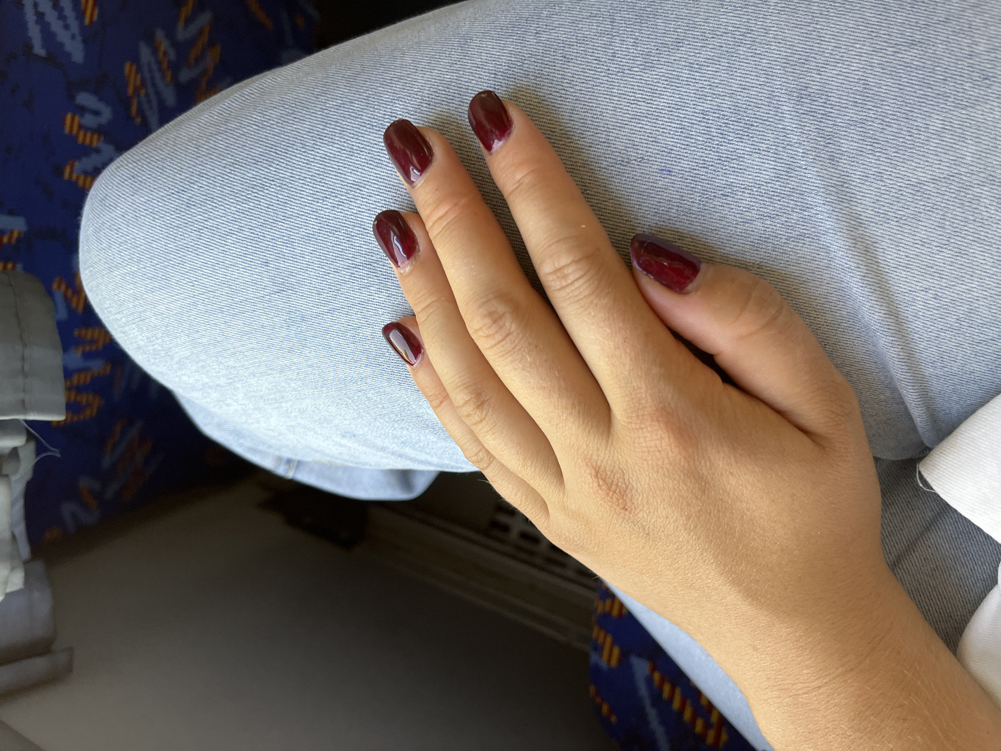 The image shows a person's hand with dark red nail polish resting on light blue denim jeans. The background includes a glimpse of a blue and orange patterned seat, possibly from a bus or train, and a dark, shadowed area. The lighting suggests an indoor setting during the day. There is no information to suggest the location is in Pastavy, Belarus.