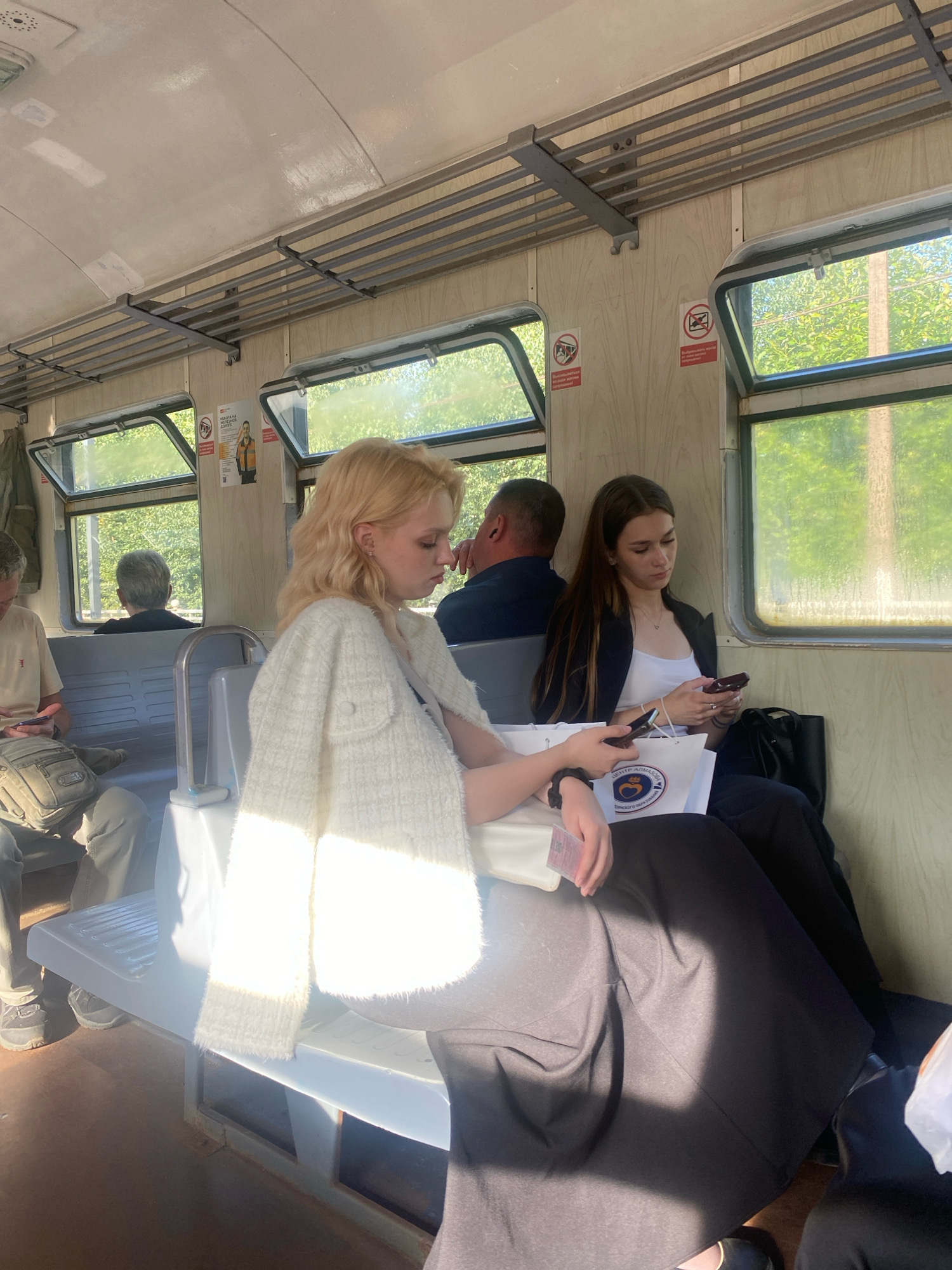 The image depicts the interior of a train carriage, likely in Saint Petersburg, Russia, during daylight. The carriage has beige wooden-paneled walls and a white ceiling with overhead luggage racks. Several passengers are seated on benches. In the foreground, a young woman with blonde wavy hair is wearing a white textured jacket over a dark garment. She is holding a phone and a small card in her hand. To her right, another young woman with long dark hair is dressed in a white top and a dark jacket, also holding a phone. Behind them, a man with short gray hair is visible from the back. On the left side of the image, a man in a light-colored shirt and khaki pants is seated, looking at his phone. A man with short dark hair is also seated further back. The windows show a view of green trees and sunlight. There are various signs on the walls, including safety or informational notices. The overall scene is of everyday travel on public transportation.