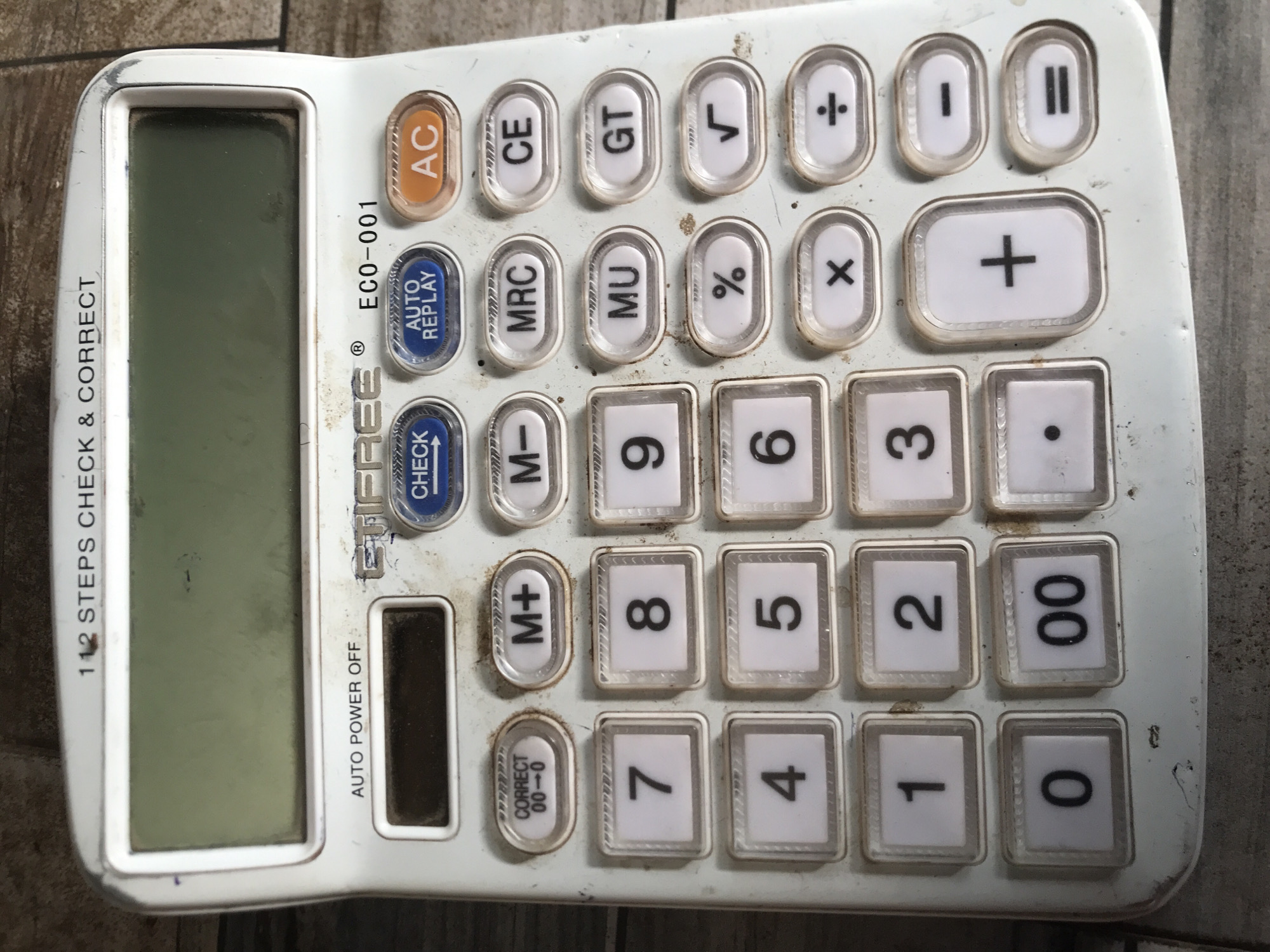 The image displays a used, white electronic calculator from a top-down perspective, positioned on a textured, possibly wooden or tiled, surface. The calculator appears well-worn, with visible dirt, grime, and minor scratches on its plastic casing and around the buttons.
On the left side of the calculator is a rectangular, dark LCD screen that is currently blank. To its right are numerous plastic keys. The top row includes an orange "AC" (All Clear) button, followed by white keys for "CE" (Clear Entry), "GT" (Grand Total), "√" (square root), and "÷" (division). The second row features a blue "AUTO REPLAY" button, then white keys for "MRC" (Memory Recall/Clear), "MU" (Mark-Up), "%" (percentage), and "x" (multiplication). The third row includes "I" (pause) and "II" (play/equals) keys, along with a large "+" (addition) key.
Numeric keys (0-9, and "00") are arranged in a standard calculator layout. Other function keys include "M+" (Memory Plus) and "M-" (Memory Minus). Below the screen, there's a blue "CHECK" button and an oval "CORRECT 00-0" button.
Visible text on the calculator body includes "112 STEPS CHECK & CORRECT" printed vertically along the left edge of the screen, "AUTO POWER OFF" below the screen, and the brand "EFIFREE®" with model number "ECO-001" printed above the "AUTO REPLAY" button. The object is static, indicating no active event or activity. The worn condition of the calculator suggests frequent use. This item is presented in the location context of Jos, Nigeria.