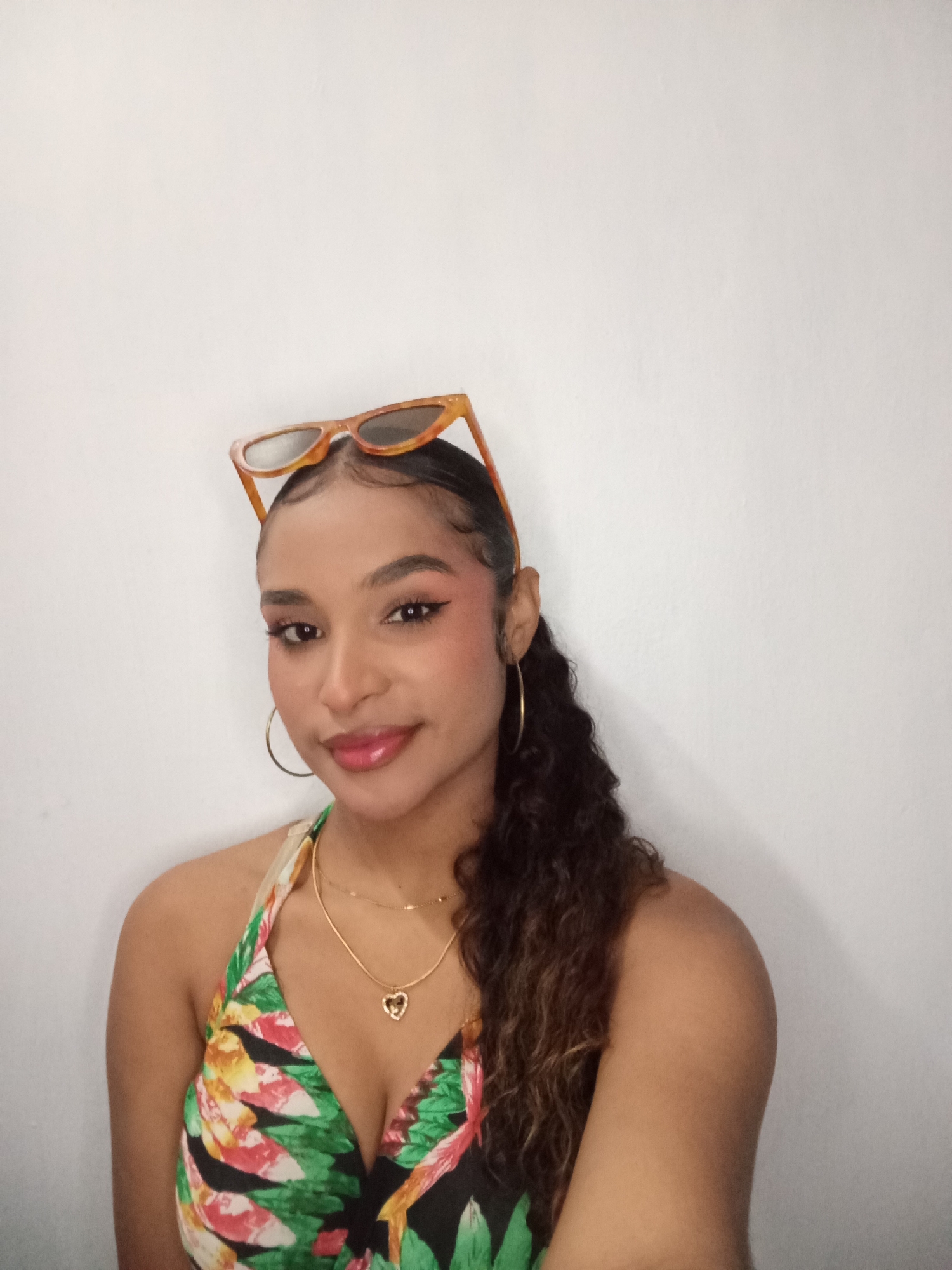 A young woman poses indoors against a plain white wall. She is wearing a floral-print halter top, gold hoop earrings, and two gold necklaces, one with a heart pendant. Sunglasses are perched on top of her head. She has curly hair pulled back into a ponytail and wears makeup, including blush, eyeliner, and lip gloss. The location, Juan Griego, Venezuela, cannot be confirmed from the image.