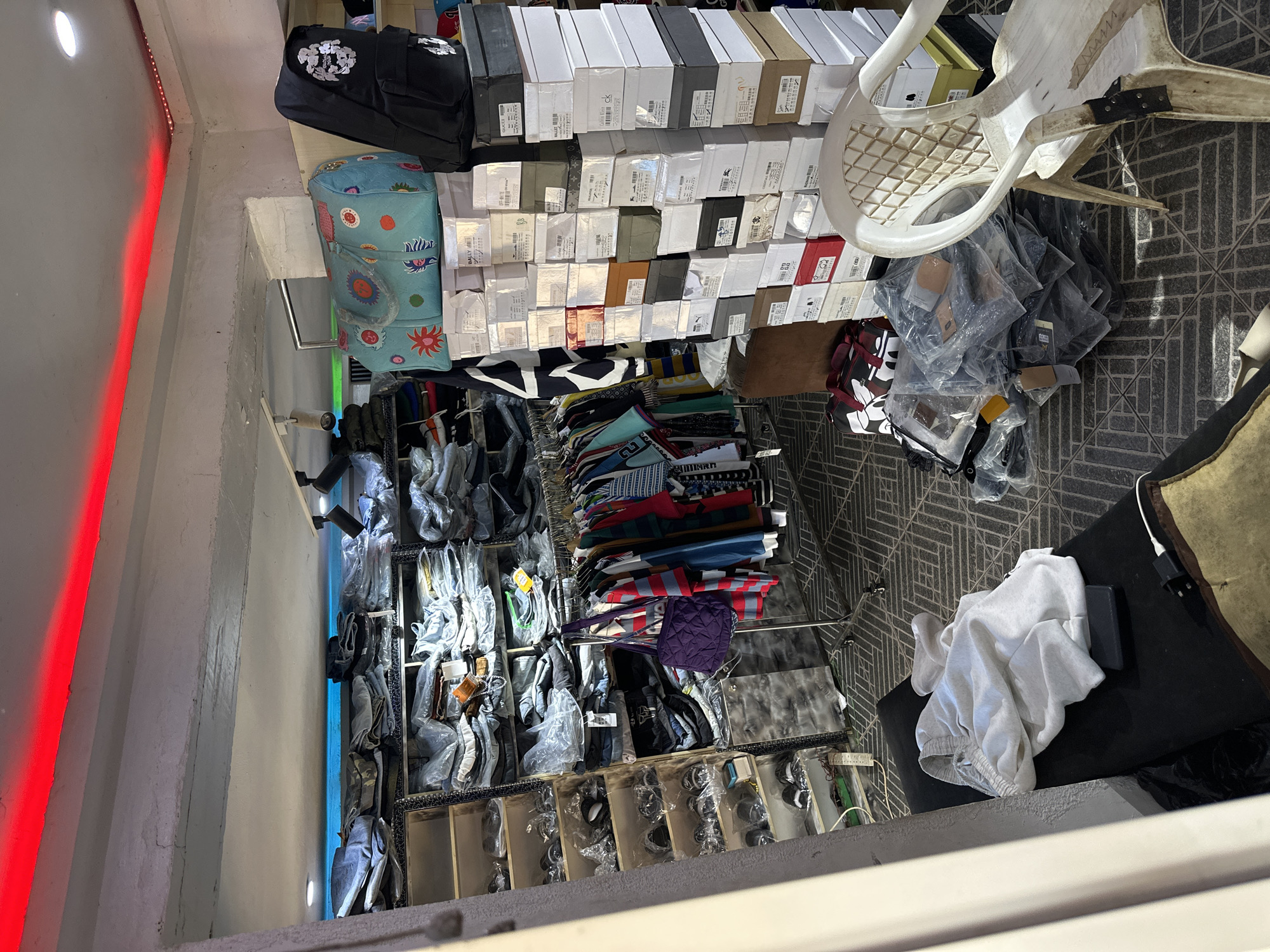 The image depicts the interior of a retail clothing store located in Bukuru, Nigeria. The store is packed with merchandise, primarily clothing items and footwear, neatly organized on shelves and racks. Various types of clothing, including jeans, shirts, and what appear to be tracksuits, are visible, some individually packaged and others hanging on display. Numerous shoeboxes are stacked high, indicating a substantial inventory of footwear. A selection of bags, including a large patterned duffel bag and a black backpack, are also present. A white plastic chair sits prominently in the foreground, adjacent to a pile of clothing and accessories. The store is illuminated by overhead lighting, with red LED strip lights running along the ceiling. The overall environment suggests a busy retail space with a wide array of products for sale. There are no people visible in the image.