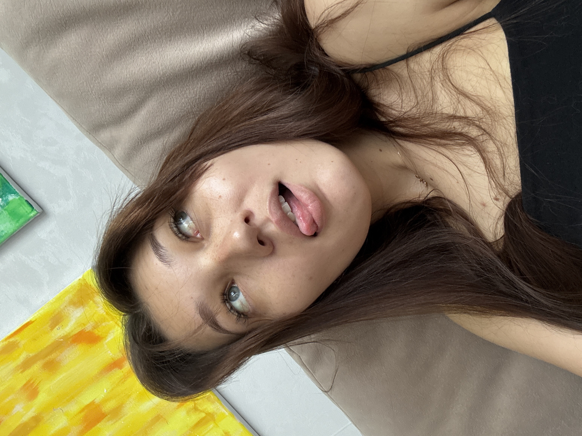 A young woman with long, brown hair and blue eyes is lying on a couch, looking up and to the right. Her tongue is sticking out slightly. She is wearing a black tank top. Behind her, a large, abstract painting with yellow and orange strokes is visible on a white wall. The couch appears to be upholstered in a light brown, textured fabric. The scene suggests an indoor setting, possibly a living room or studio space. The location is not specified as Novokuznetsk, Russia.