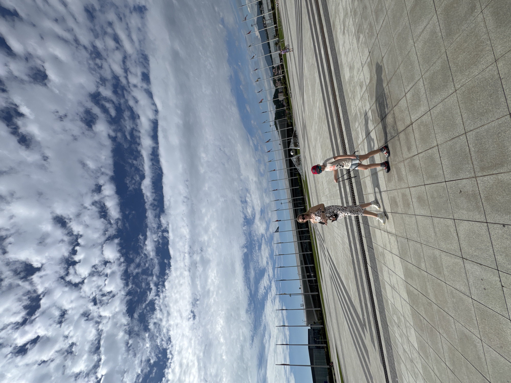 The image shows an outdoor scene in the city of Sirius, Russia. The sky is visible with blue patches and white clouds. A paved area is present, with two individuals standing on it. One individual is a child wearing a red helmet, a t-shirt, and shorts, while the other is an adult in a patterned dress and sunglasses. There are flagpoles lined up in the middle ground, and a body of water and possibly other buildings are visible in the background.