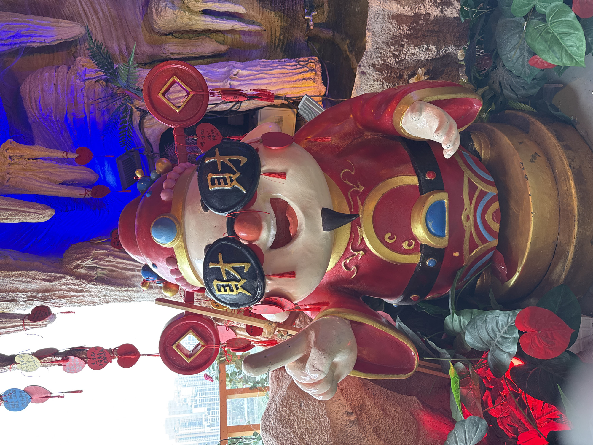 A close-up, medium-angle shot presents a colorful statue of a Chinese figure, likely a representation of a god or deity, set against a backdrop of artificial rock formations and greenery. The statue is predominantly red and white, with gold accents highlighting the details of the clothing and features. The figure is wearing round, black sunglasses with Chinese characters inscribed on them. The figure has a wide, toothy grin. Coins with square holes are attached to the statue, and a collection of colored tags with Chinese writing hang from the figure. The setting appears to be an indoor display or shrine. In the background, there is a glimpse of tall buildings, suggesting an urban location. Based on this, the location is determined to be within Yuzhong District, China. The scene seems to be a combination of traditional religious symbolism and contemporary urban surroundings.