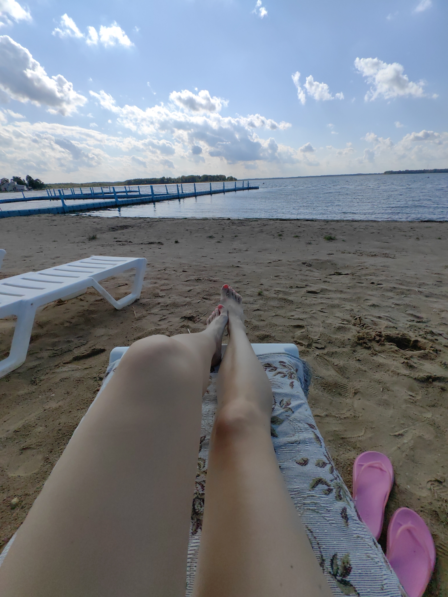 In Myadzyel, Belarus, on a sandy beach next to a calm body of water, two white lounge chairs are positioned on the sand. A person's legs are visible, resting on one of the lounge chairs, with pink flip-flops to the right. The sky above is blue with scattered white clouds. In the distance, a blue pier extends into the water, and a treeline is visible on the far shore.