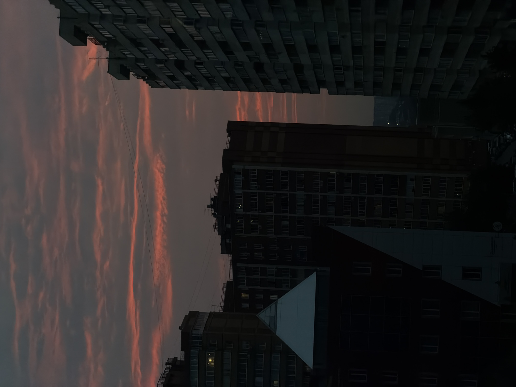 This is a photograph of the sky at sunset, seen between tall apartment buildings in Novosibirsk, Russia. The sky is a deep grey, with streaks of vibrant orange and pink clouds stretching vertically across the frame, suggesting they are illuminated by the setting sun. The clouds appear wispy and elongated, creating a dramatic and somewhat surreal effect.
The foreground is dominated by the dark silhouettes of several multi-story apartment buildings. One building, at the top of the frame, is a long, uniform structure with many windows. Below it, another building shows a darker facade with a band of yellowish windows. Further down, a building with a striking triangular section covered in a light-colored material juts out. The windows of these buildings are mostly dark, with a few showing faint lights, indicating that it is dusk and people are home. The composition is such that the buildings frame the colorful sky, creating a stark contrast between the urban environment and the natural beauty of the sunset.
The overall mood is one of quiet twilight, with the vibrant sky offering a moment of beauty amidst the imposing concrete structures. The time of day is clearly sunset, and the weather appears clear apart from the cloud formations. There is no visible text in the image. The perspective is looking upwards, implying the photographer is likely on a lower floor or ground level, observing the sky from between the buildings.