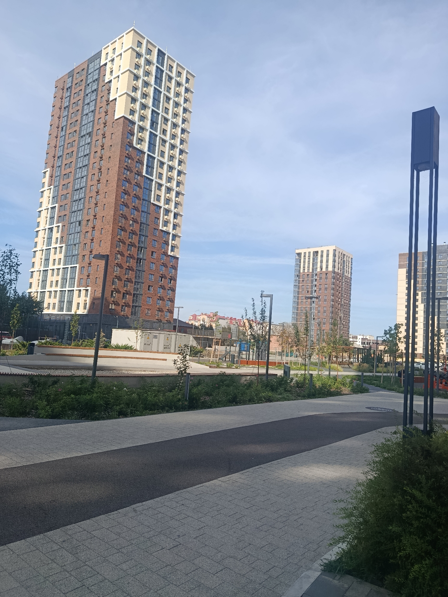 The image depicts a modern urban landscape with several high-rise buildings under a clear blue sky. In the foreground, a paved walkway curves through a landscaped area with greenery and trees. To the left, a tall, slender building with a brick facade and cream-colored accents dominates the view. Further in the distance, two other similar high-rise buildings are visible, along with some lower structures and construction cranes, suggesting ongoing development. A dark, rectangular monument or light fixture stands to the right of the frame. The overall scene conveys a sense of new construction and residential development in a city. The specific location of Astrakhan, Russia is not visually identifiable within the image itself.
