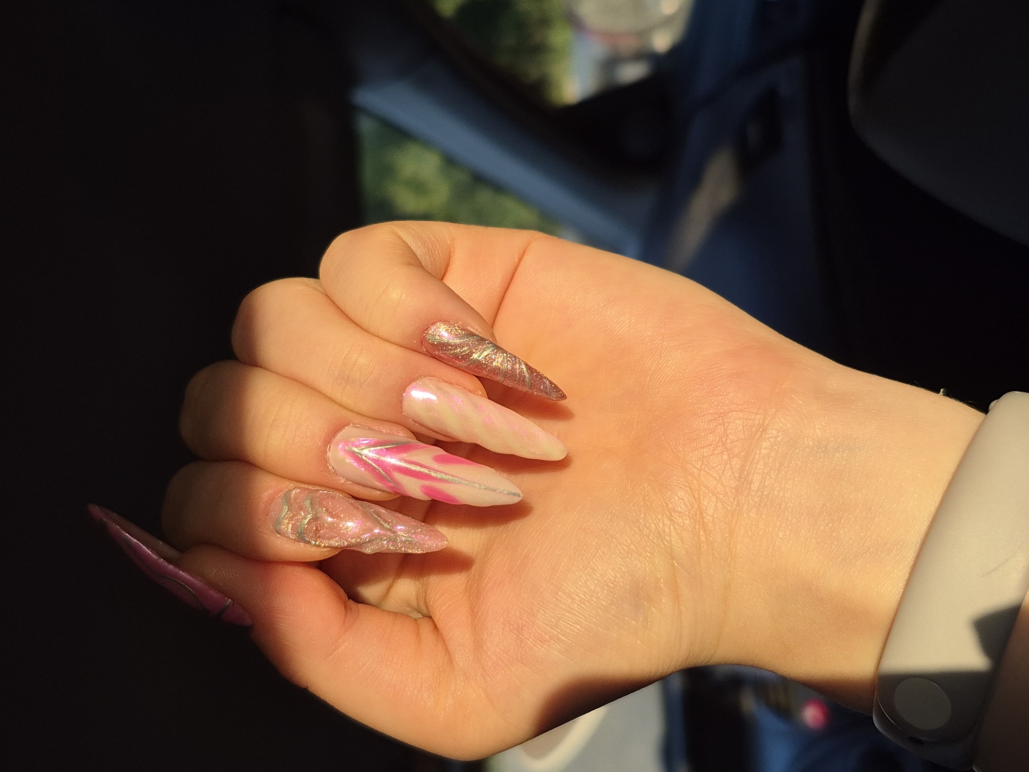 The image shows a close-up of a person's hand, which is partially illuminated by sunlight casting shadows. The hand has long, stiletto-shaped acrylic nails. The nails are adorned with a marble or marbled effect in shades of pink, white, and silver, with some having a glitter or metallic finish. The person is wearing a light-colored band, possibly a watch or fitness tracker, on their wrist. The background is dark and out of focus, with some blurred greenery and what appears to be the interior of a vehicle, suggesting the person might be in a car. The lighting indicates it is daytime, likely a sunny day, given the strong shadows. The image focuses on the detailed nail art and the texture of the skin. No text is visible in the image. The overall impression is a focus on personal grooming and style.