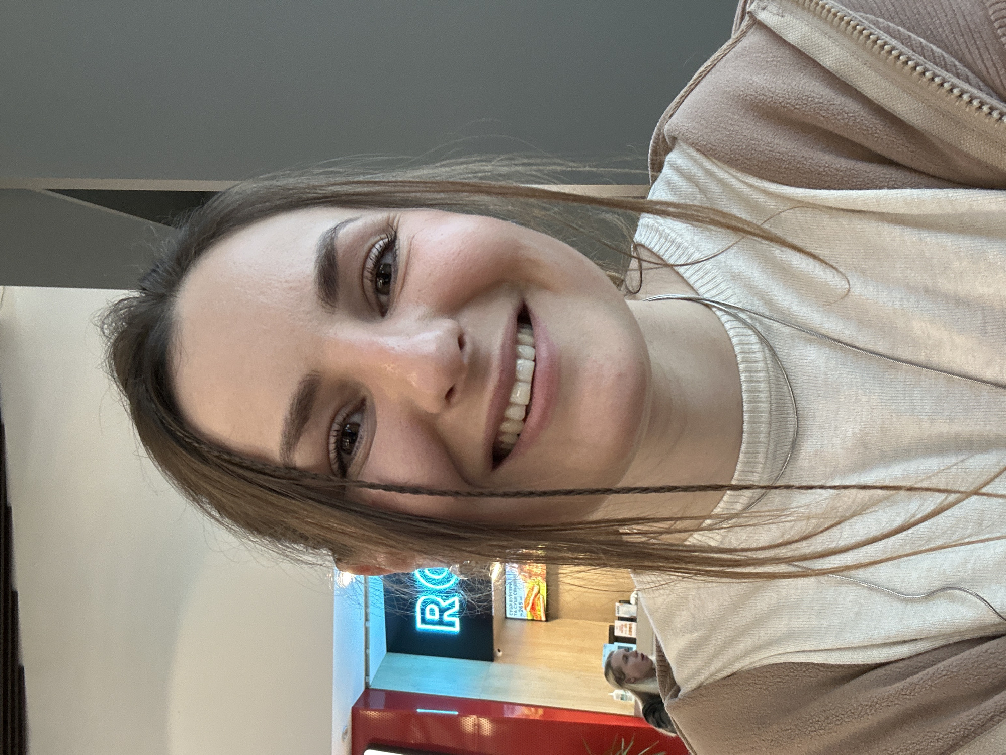 A young woman with brown hair and a warm smile is positioned in the foreground, wearing a white shirt and a brown jacket. The background reveals what appears to be a retail environment, evidenced by the presence of a red and white counter and a brightly lit, blue neon sign with the letters "RO." A reflection of another person with blonde hair is visible in the background. The location is in the city of Unknown, Unknown.