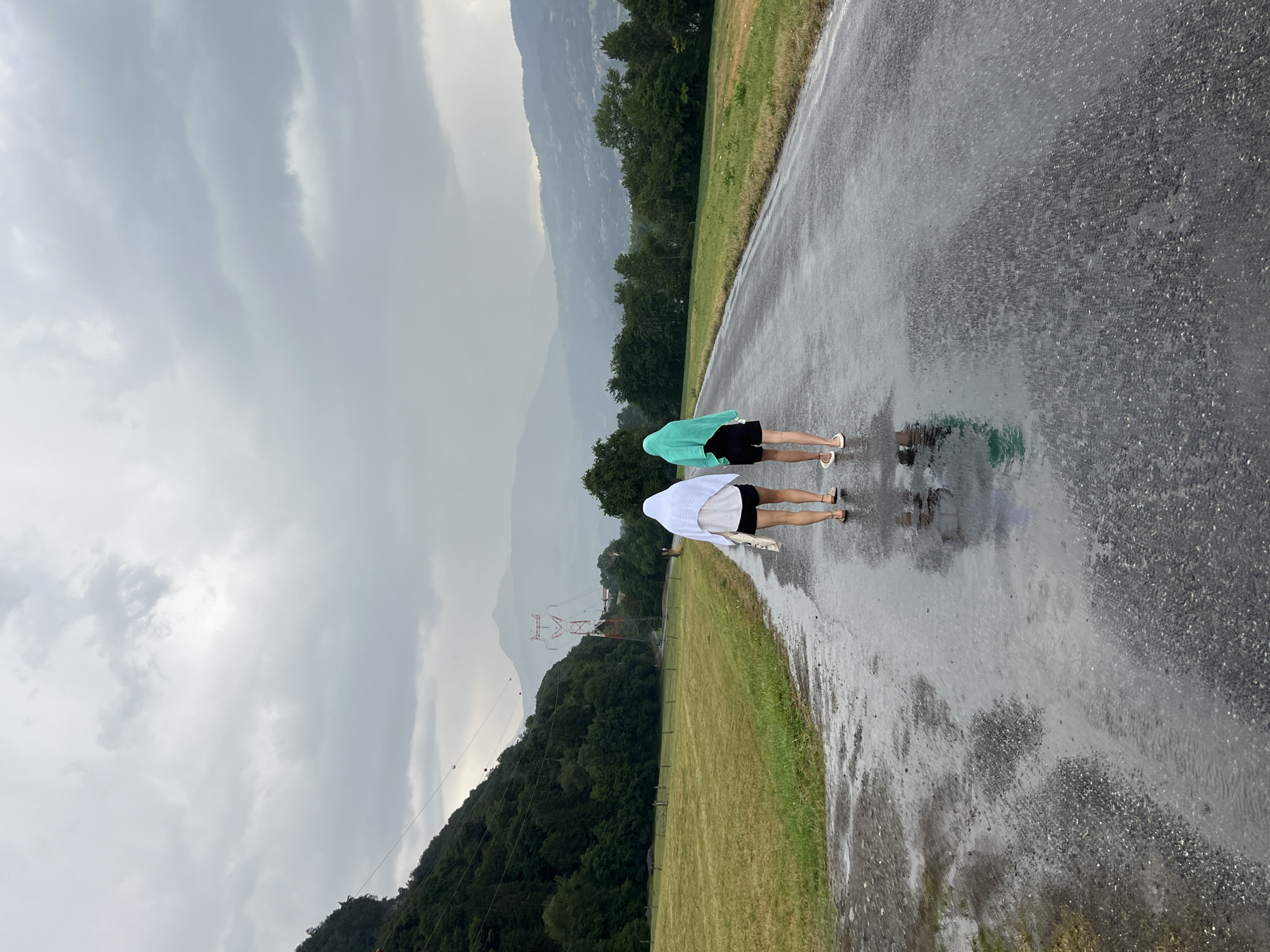 Two individuals, viewed from behind, walk away from the camera on a wet asphalt road. The person on the left wears a white towel draped over their head and shoulders, black shorts, and sandals. They carry a light-colored tote bag in their left hand. The person on the right wears a turquoise towel over their head and shoulders, black shorts, and sandals. The road is wet with visible puddles and reflections, indicating rain. To the left of the road is a grassy field, bordered by a dense line of green trees. In the background, a steep, tree-covered mountain slopes upwards. Further in the distance, more rolling hills and mountains are visible under a heavily overcast, gray sky. A large red and white electrical pylon stands in the middle ground. The scene appears to be a rural or semi-rural area, likely in or near Sallanches, France, given the mountainous landscape.