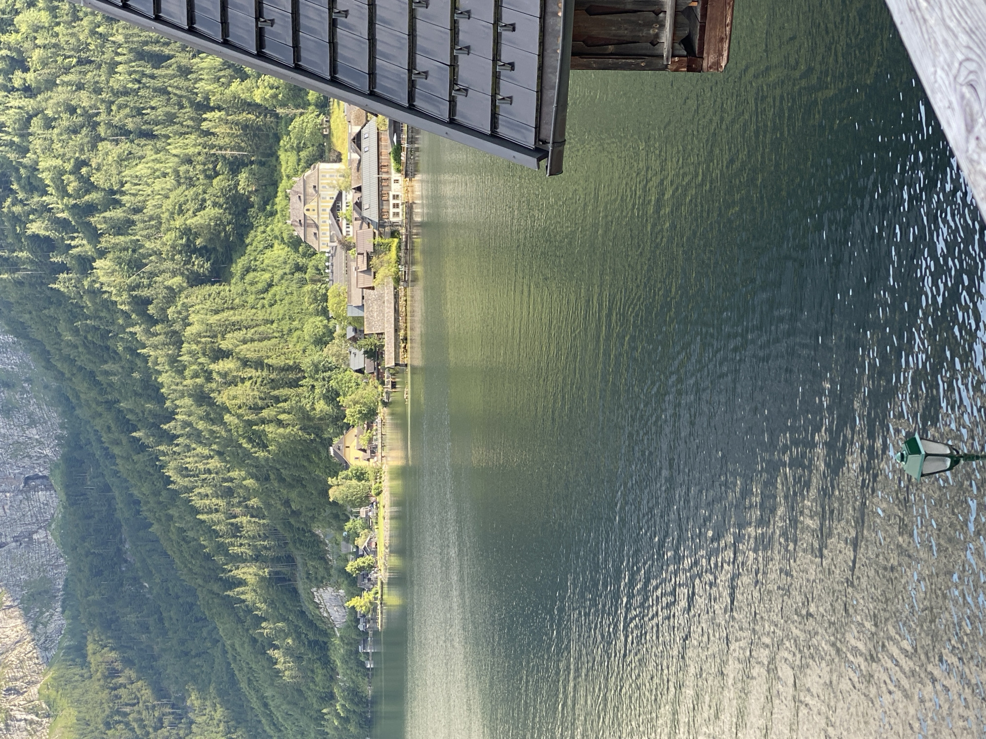 The image depicts a scenic view of Hallstatt, Austria, a UNESCO World Heritage site. The foreground is dominated by a body of dark, rippling water, reflecting the light and the surrounding landscape. In the lower right corner, a portion of a wooden structure with a dark, tiled roof is visible, angled towards the water. A green-topped lamppost stands on a wooden railing in the lower center of the frame, also positioned near the water's edge.
Across the water, the town of Hallstatt is nestled along a narrow strip of land. Numerous traditional Austrian buildings with characteristic peaked roofs are visible, painted in various muted colors, including a prominent yellow building. These structures are situated at the base of a steep, densely forested mountain that rises dramatically behind the town. The mountain is covered in a thick carpet of green trees, with lighter grey rock formations visible at its summit. The scene is bathed in natural light, suggesting it was taken during daytime, likely in the late afternoon given the angle of the shadows and the warm tone of the light. There are no people or animals visible in the image.