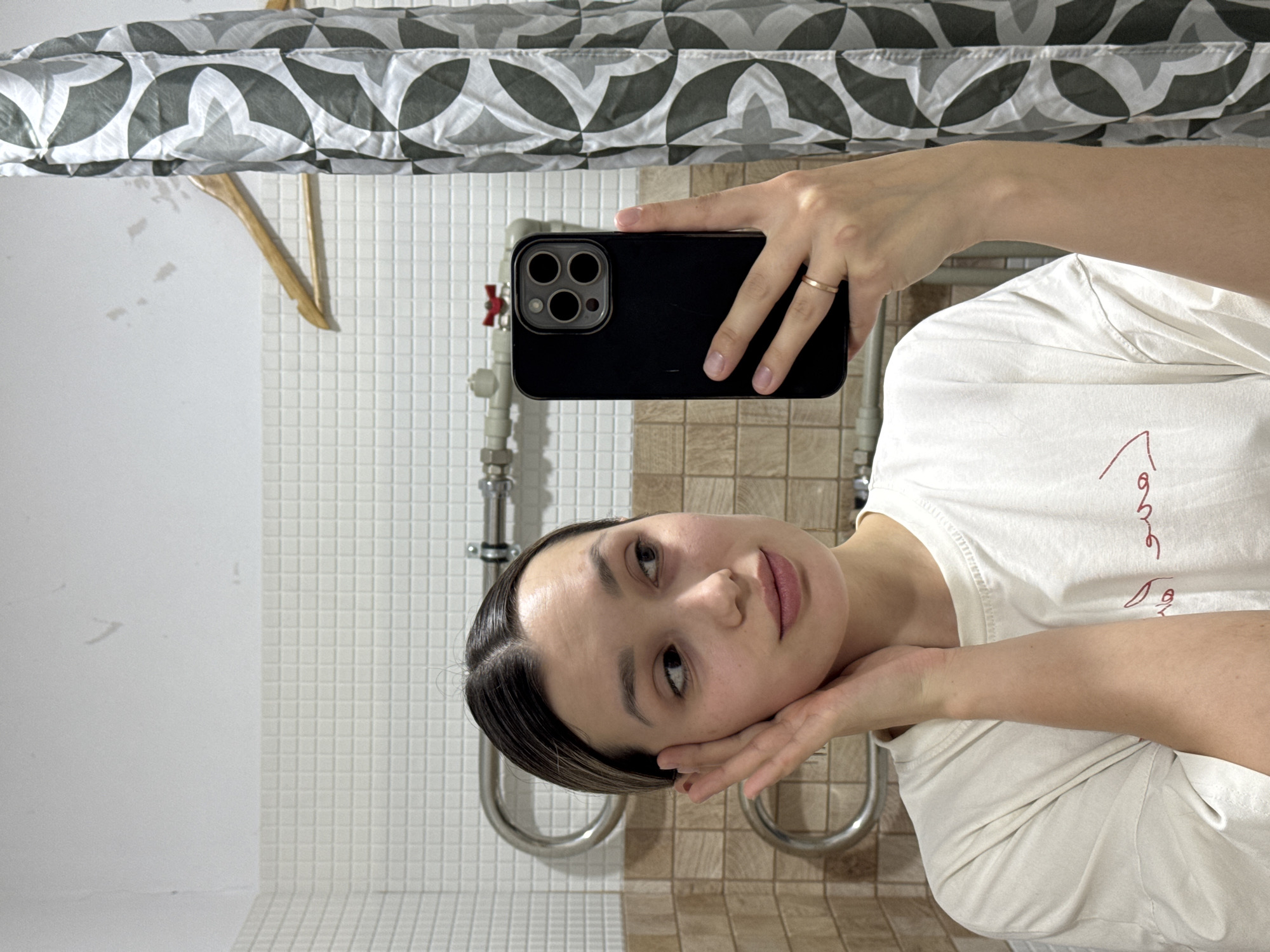 A rotated eye-level selfie shows a fair-skinned woman with dark hair in a white t-shirt holding up a black phone in a bathroom. She has a gold ring on her finger. The background includes a shower curtain with a leaf pattern and white and brown tiled walls. A wooden hanger is also visible.