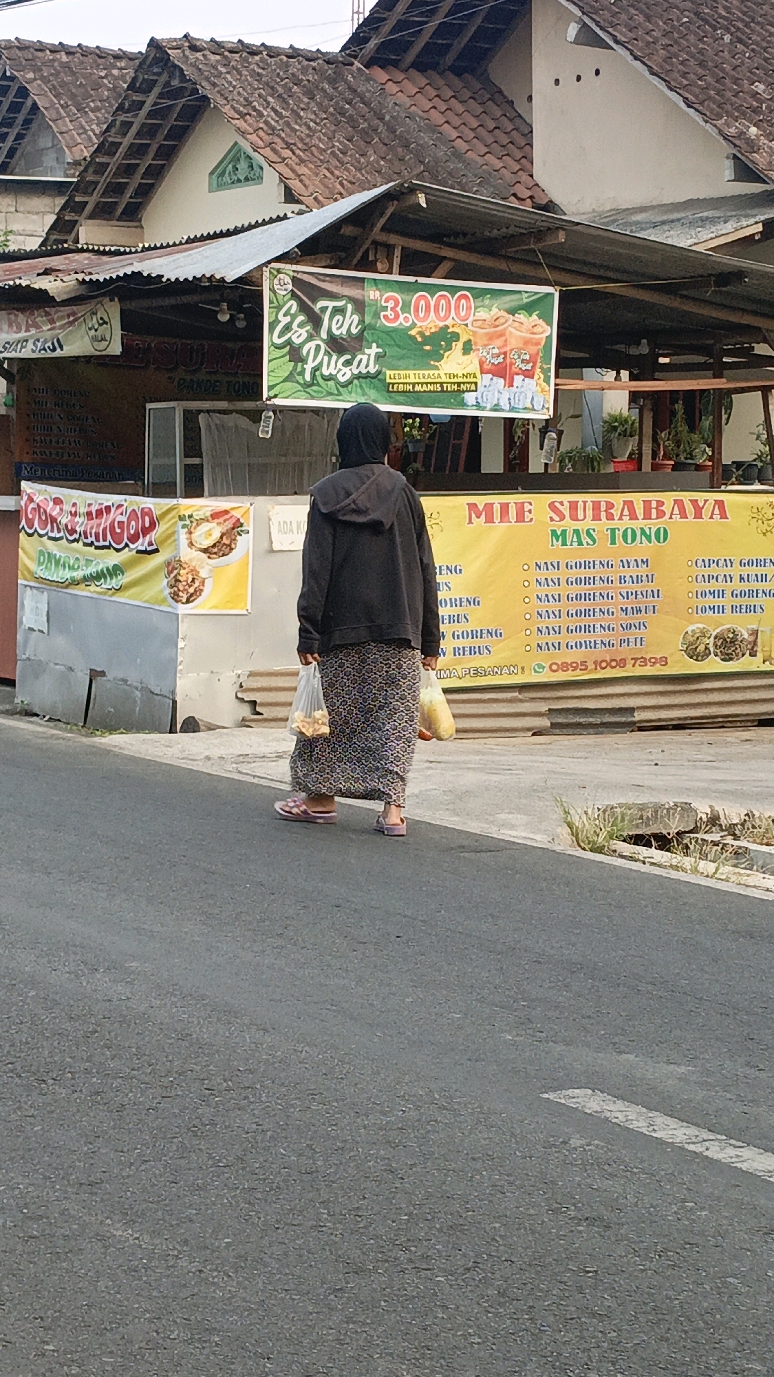 The image shows a street scene in Salatiga, Indonesia. A woman wearing a dark hijab, a dark hooded jacket, and a patterned skirt walks away from the camera on a paved road. She carries two plastic bags, one in her right hand and another in her left. To her right, a yellow banner with text in Indonesian reads "MIE SURABAYA MAS TONO," listing various noodle dishes and a phone number. To her left, a green banner advertises "Es Teh Pusat" for Rp 3,000. Behind the banners, buildings with tiled roofs are visible. The setting appears to be a commercial area, likely a street with food vendors.