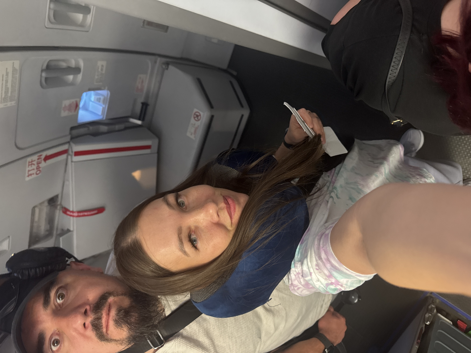 A medium shot depicts a woman with long brown hair, wearing a blue neck pillow and a white tie-dye top. She is sitting on what appears to be an airplane seat and looking directly at the camera. In the lower left corner of the frame, a man with a dark beard and headphones is visible. The background consists of the interior of an airplane, including an emergency exit door. The specific location and city are unknown.