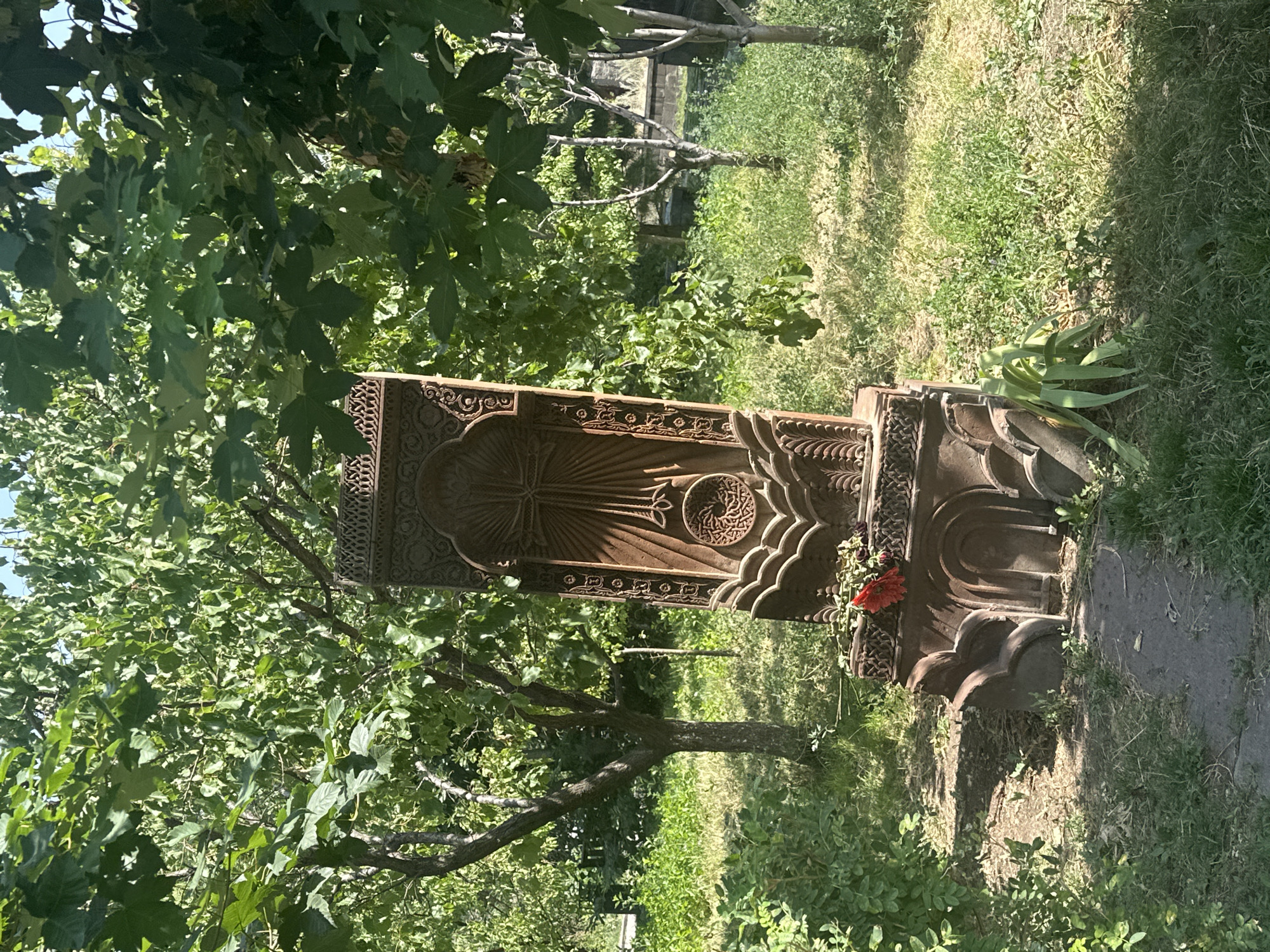 An ancient Armenian khachkar, a carved stone cross, stands in a garden or cemetery surrounded by lush greenery and trees. The khachkar is ornately carved with a central cross surrounded by radiating lines and a decorative circular motif at the base, all set within a tall, rectangular stone slab. A few red flowers lie at the base of the khachkar, suggesting a recent tribute. The scene is bathed in sunlight, with dappled light filtering through the leaves of the overhanging trees, creating a peaceful and somewhat somber atmosphere. The overall impression is one of history and reverence in a natural setting. This khachkar is located in the Marmashen Monastery complex, near the city of Gyumri, Armenia.
