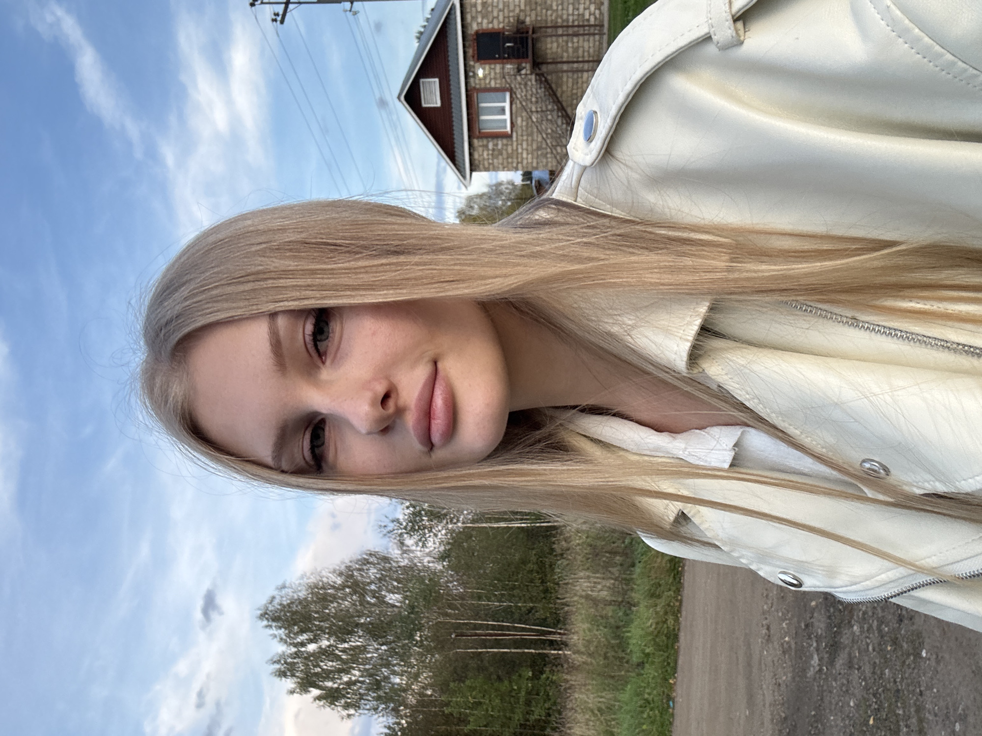 In Комарихинский, Russia, a young Caucasian woman with long, straight blonde hair is captured in a selfie. She is wearing a cream-colored leather jacket over a white collared shirt. Her gaze is directed towards the camera, and her lips are slightly parted. The background features a dirt road, a line of trees and foliage, a small brick building with a red roof and an external staircase, and a sky with scattered clouds. A utility pole with wires is also visible. The scene appears to be set in a rural or semi-rural environment, possibly during the late afternoon or early evening given the lighting conditions.